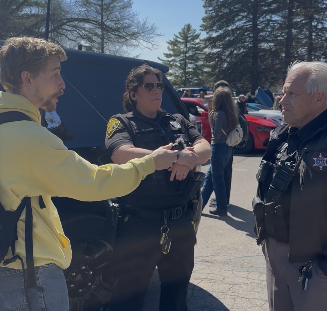 A young man in a yellow hoodie shakes hands with a police officer outdoors, with trees and parked cars in the background.