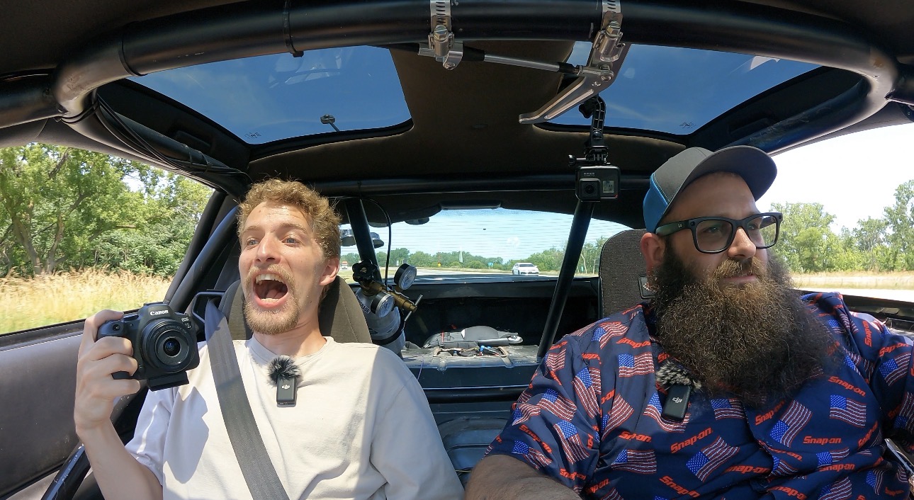 Two men sitting in a car, one smiling and holding a camera, the other wearing glasses and a hat, with a landscape outside.