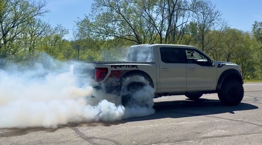 Pickup truck performing a burnout, producing thick white smoke from rear tires, on a paved road with trees in background.