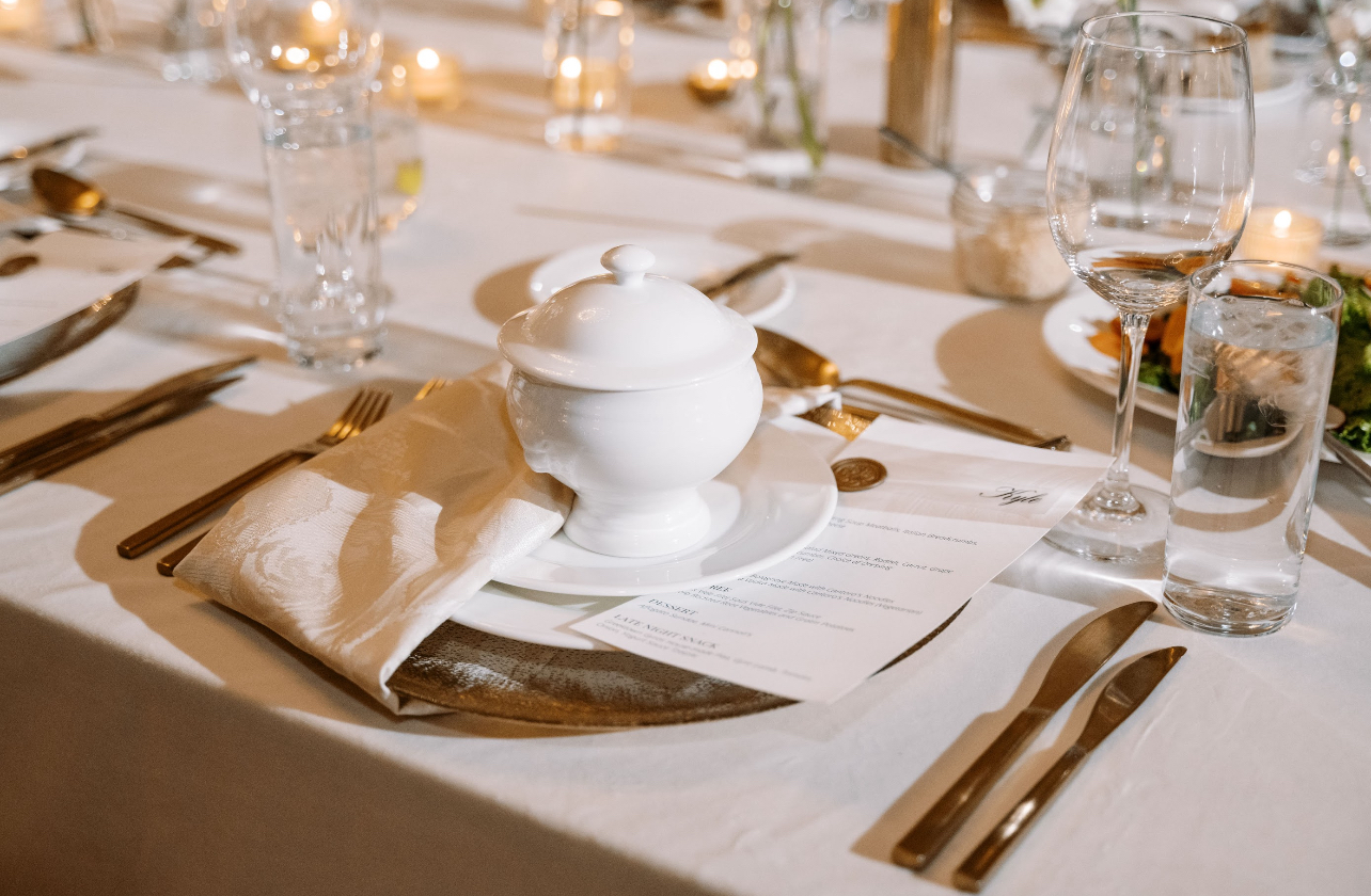 Elegant table setting with a white ceramic dish, napkin, cutlery, glasses, and a menu on a white tablecloth.