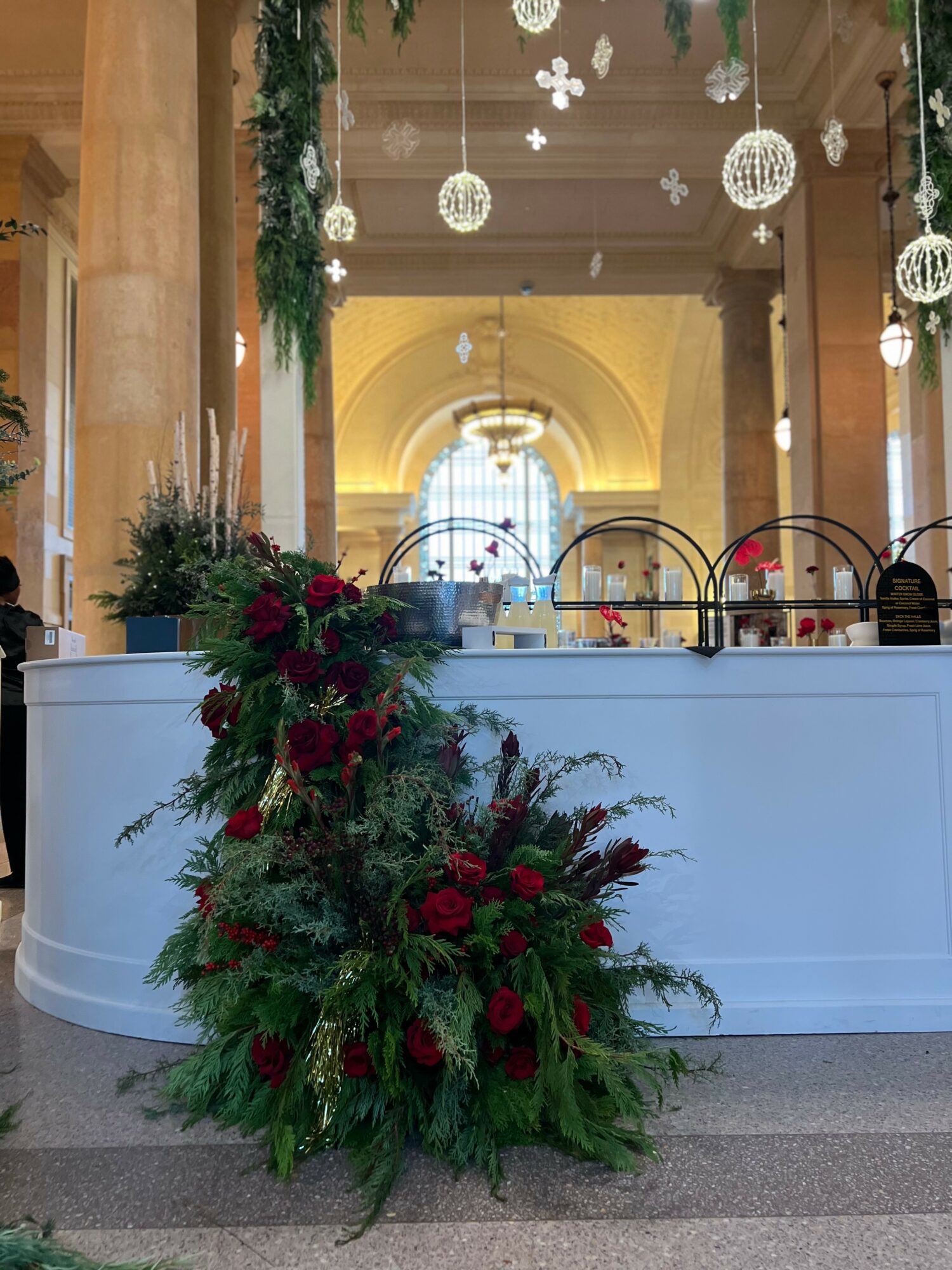 Decorative floral arrangement with red and green foliage in front of a white counter, inside a decorated hall with hanging ornaments.