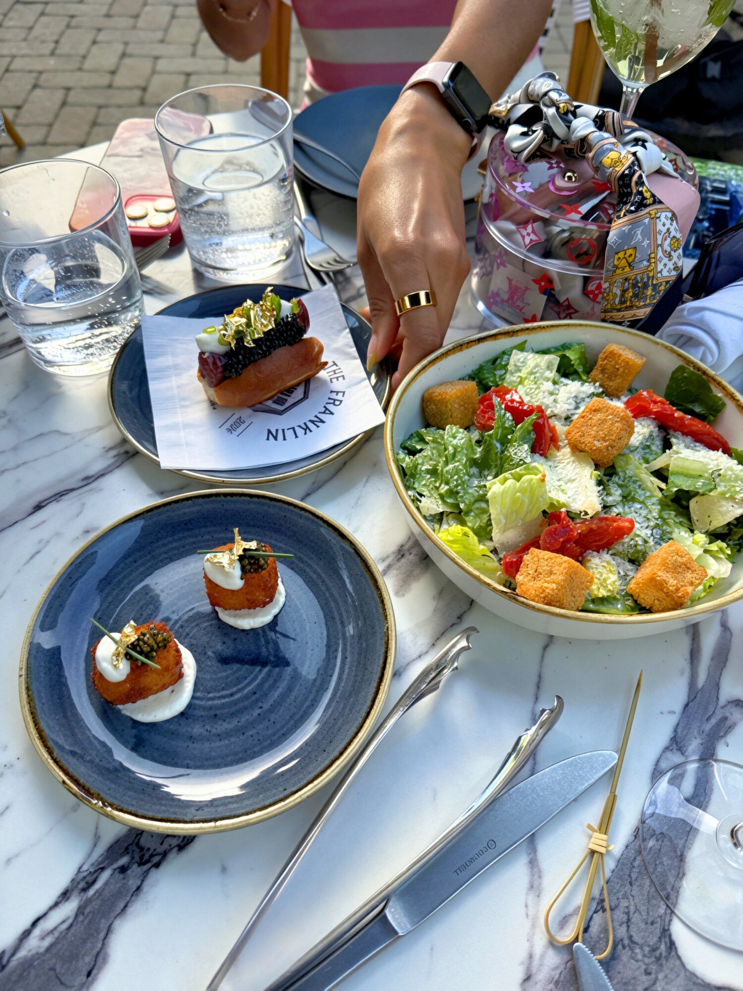 Table with salad, two small desserts, glasses of water, and hands of people at outdoor dining setting.