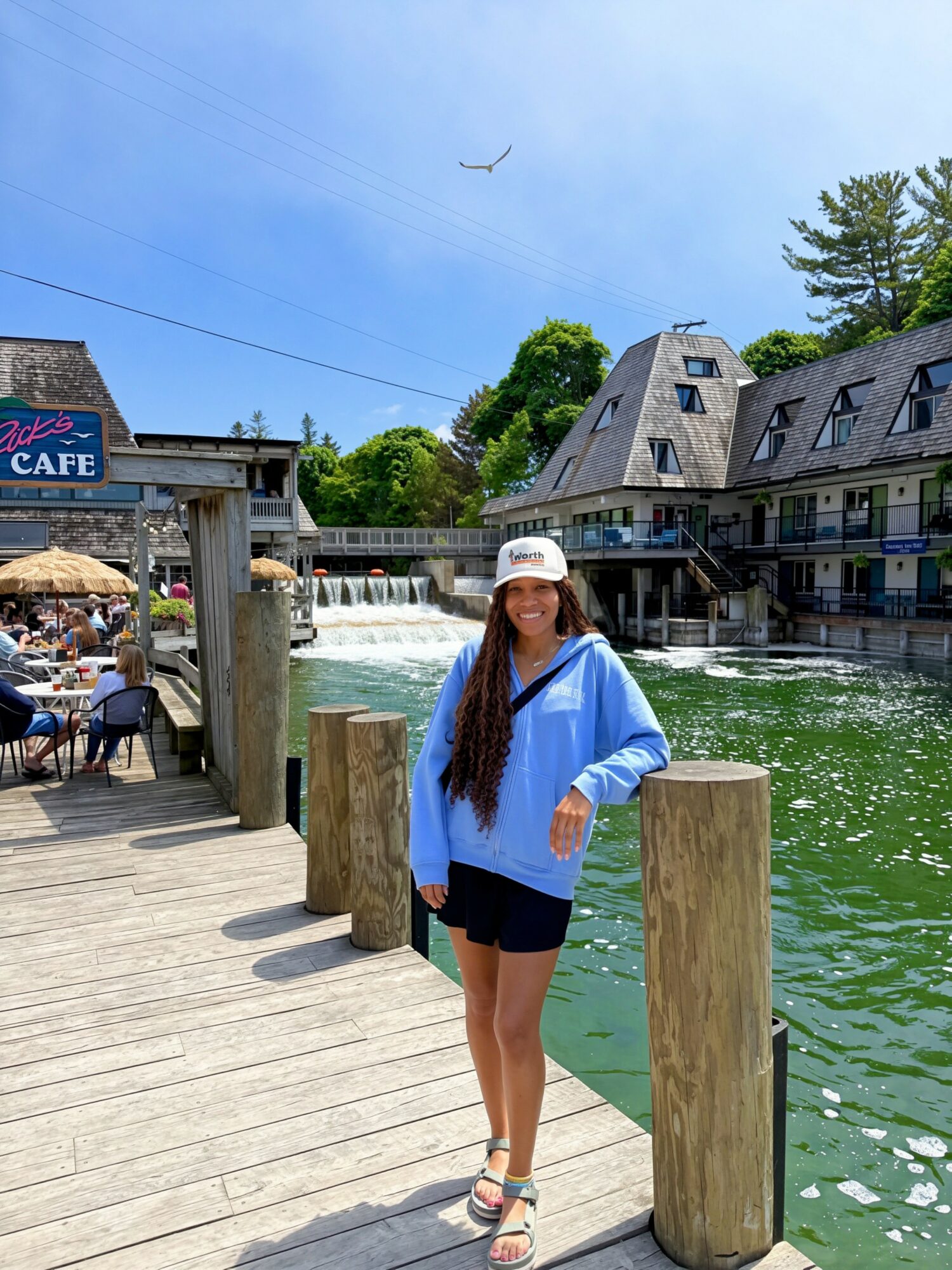 Woman in blue jacket and hat standing on a wooden dock near water, buildings, and trees, with a waterfall in the background.