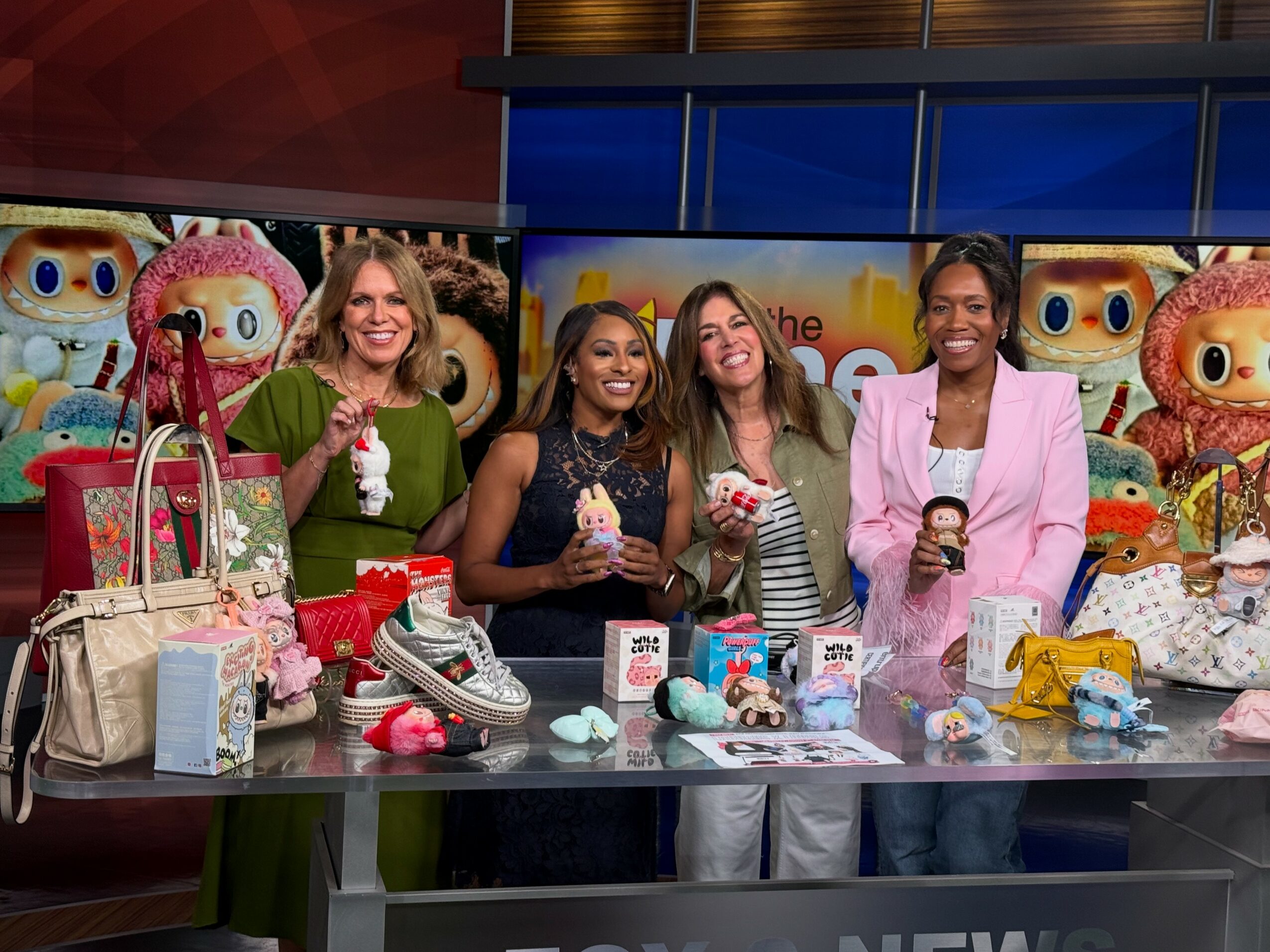 Four women stand behind a table with toys and merchandise, smiling in a colorful studio setting.