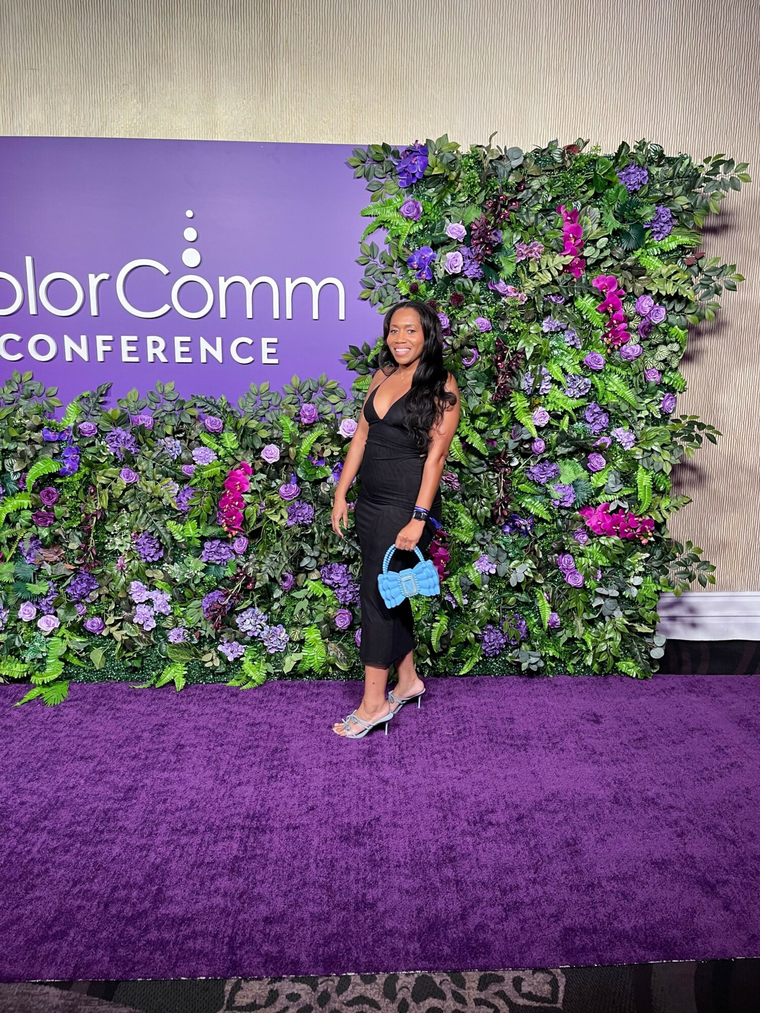 Woman in black dress standing next to a purple floral backdrop at a conference, with purple carpeted floor.