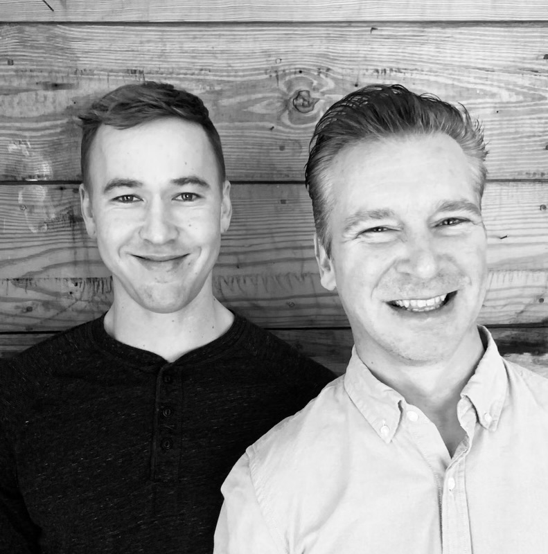 Two men smiling in front of a wooden wall, one wearing a dark shirt, the other a light shirt, in black and white.