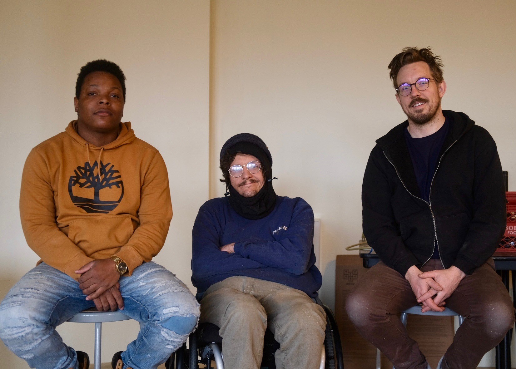 Three people sitting against a plain wall, smiling, with casual clothing and glasses, in a relaxed setting.