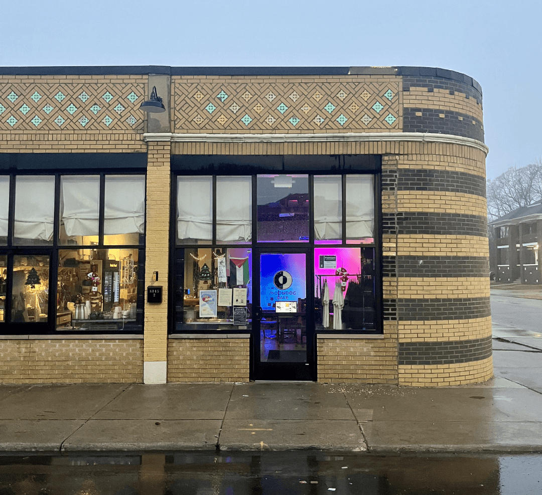 Storefront with large glass windows and a door, brick exterior, and a decorative upper facade, on a wet sidewalk.