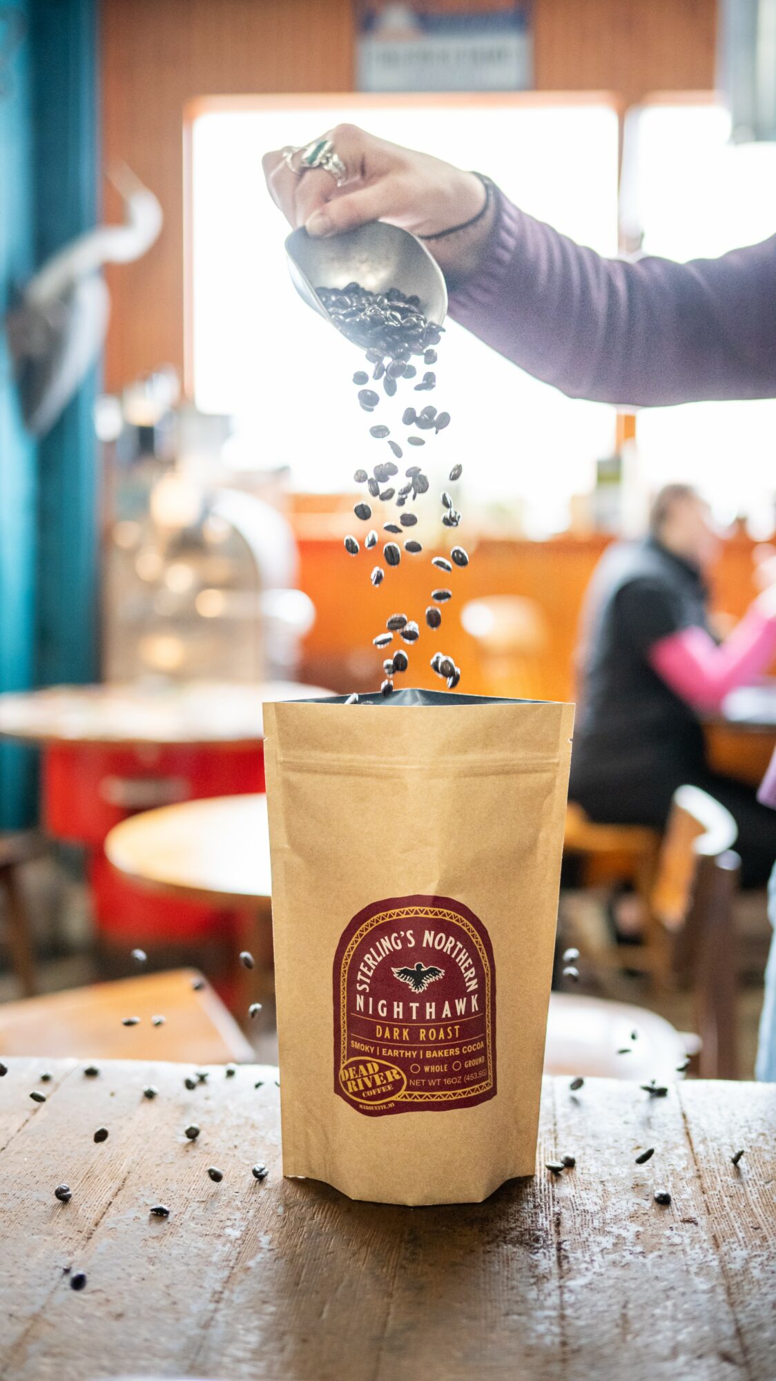 Person pouring coffee beans into a paper bag in a cafe, with people sitting in the background.