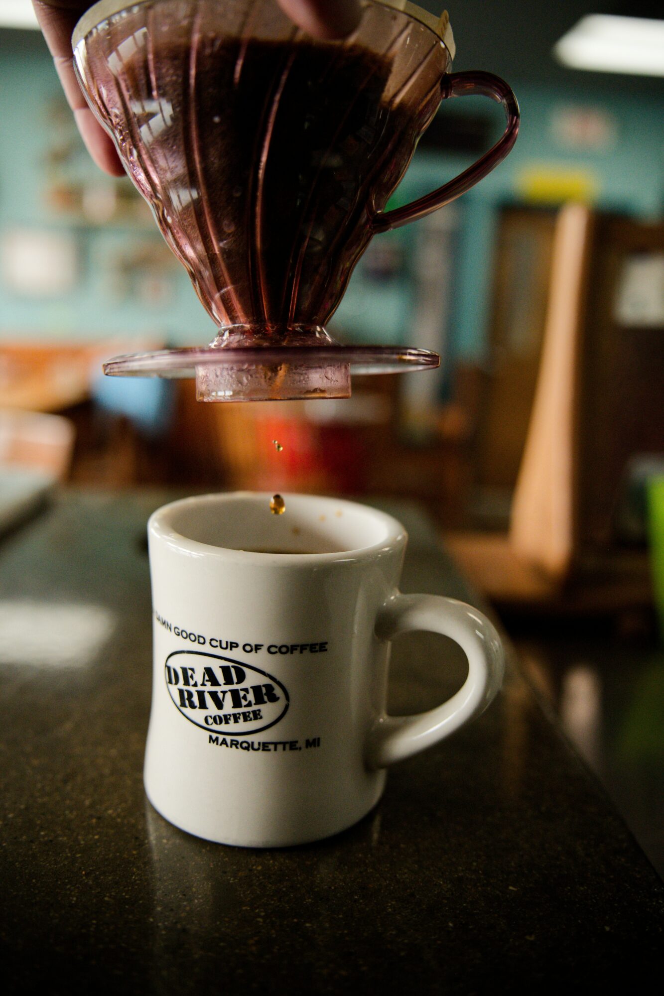 Coffee filter pouring hot coffee into a white mug with 'Dead River' logo, on a dark surface, blurred background.