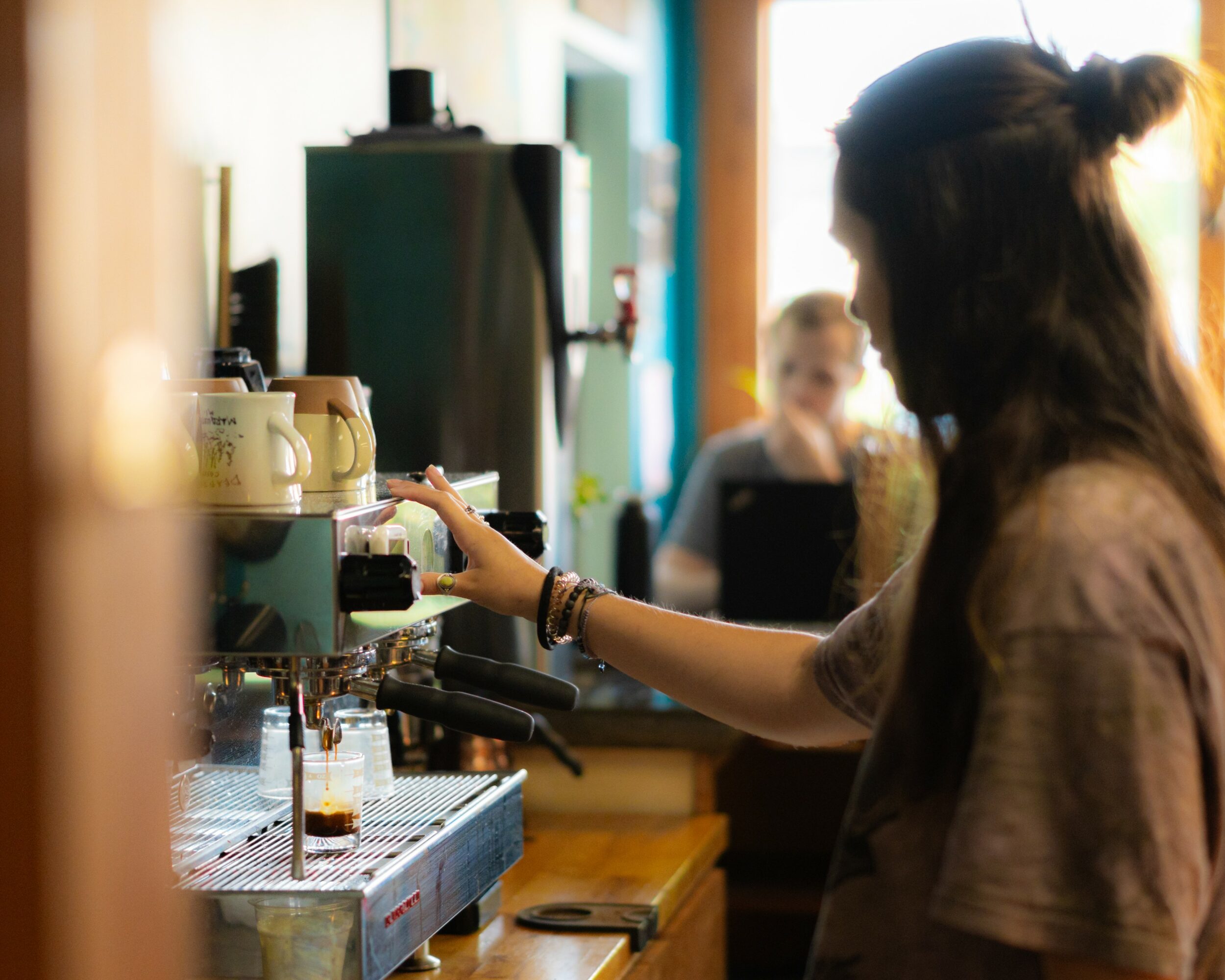 Person operating coffee machine in a cafe with a barista in the background, warm lighting, and coffee cups on the counter.