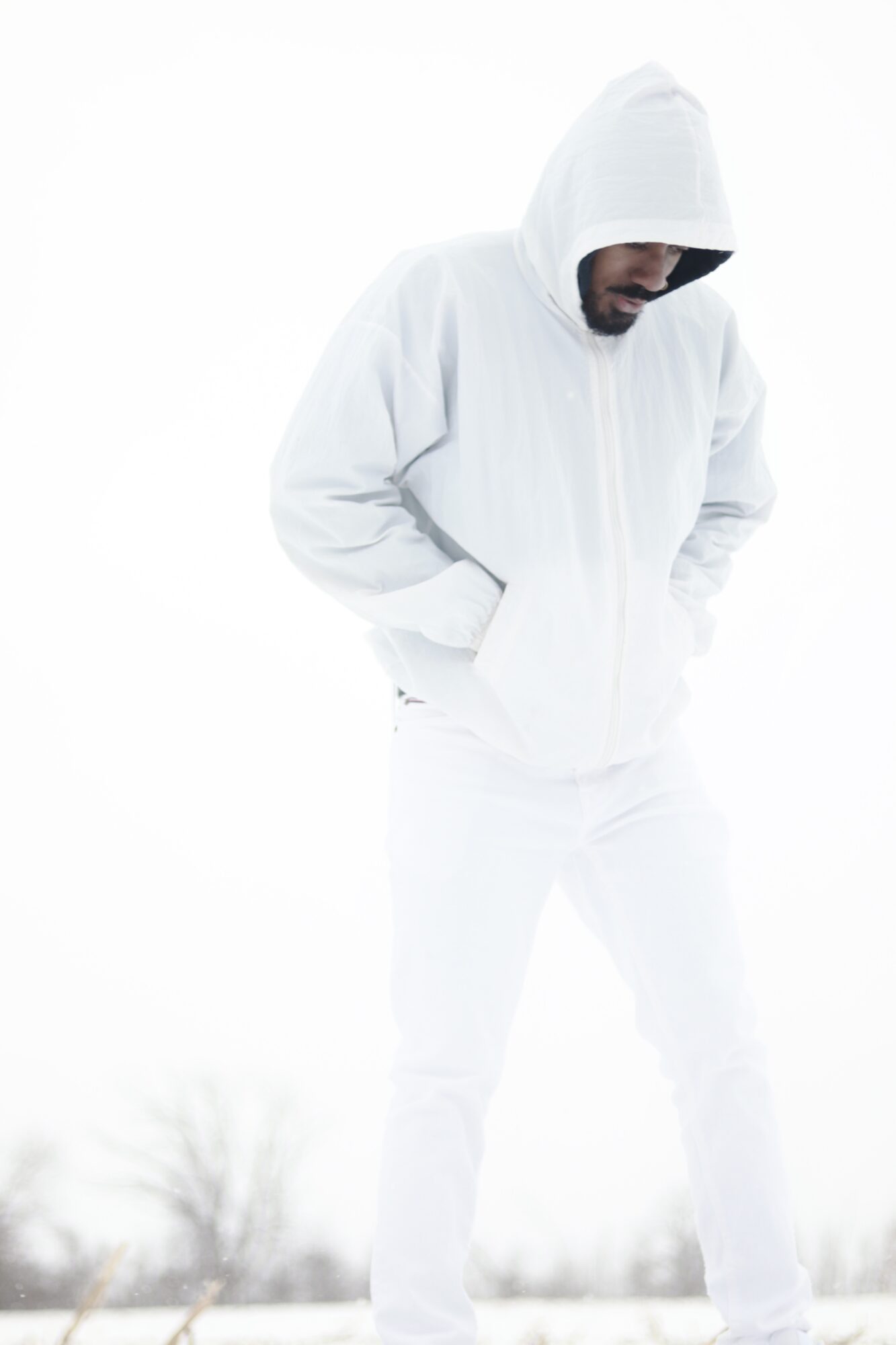 Person in white hoodie and pants looking down with hands in pockets, outdoors in winter, overcast sky.