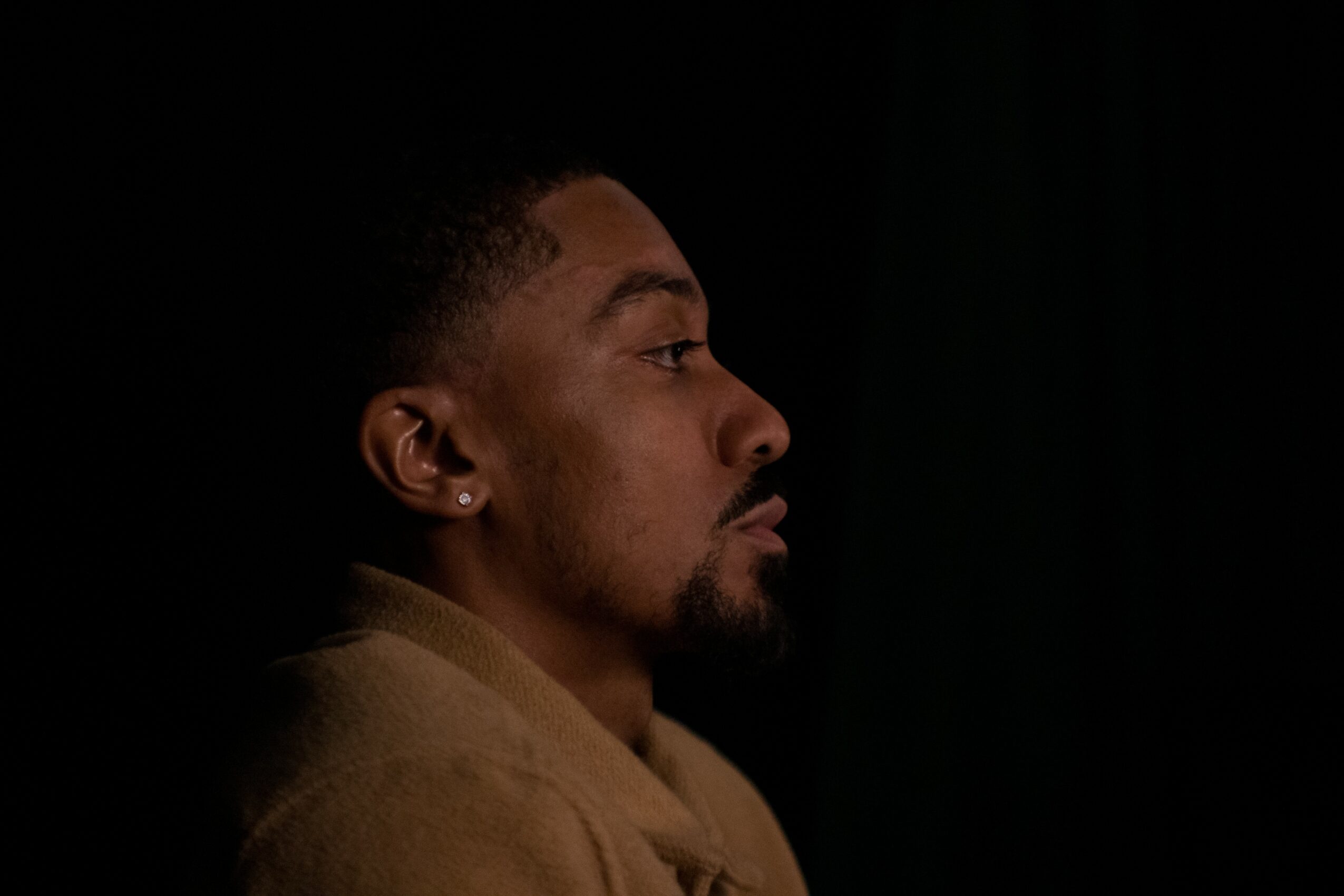 Profile of a man with a beard and earrings, facing right, against a dark background.