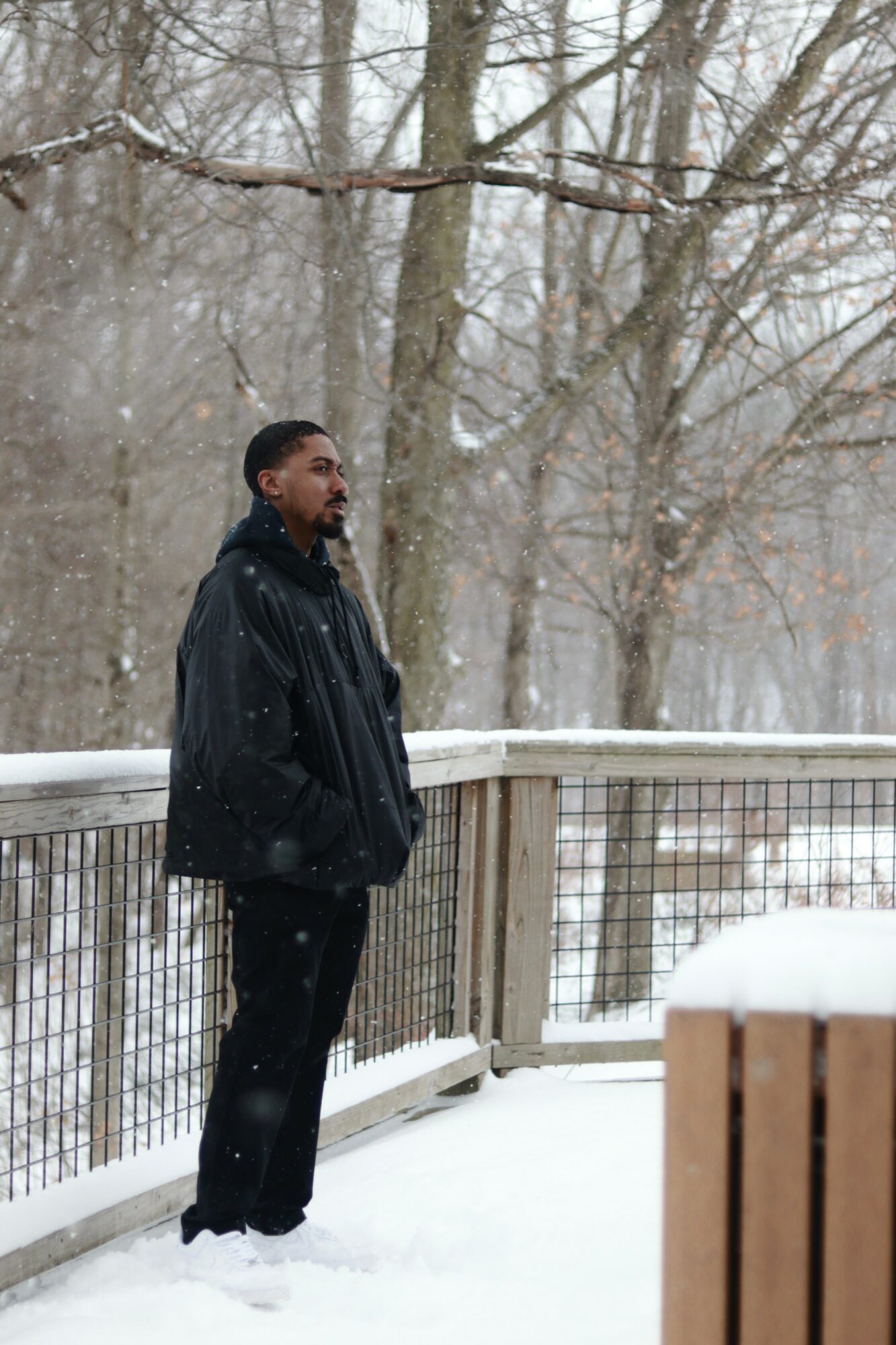 Man standing on snowy deck with railing, trees in background, snow falling, winter scene.