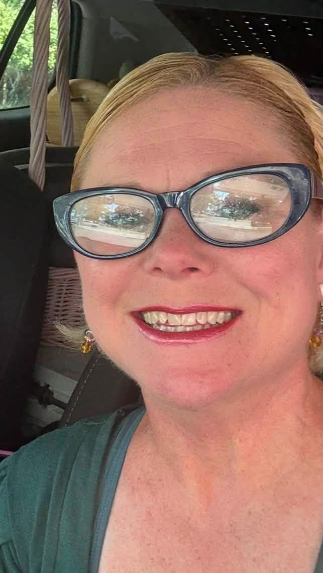 Woman with glasses smiling, sitting in a vehicle, sunlight reflecting on glasses, blonde hair, wearing a green top.