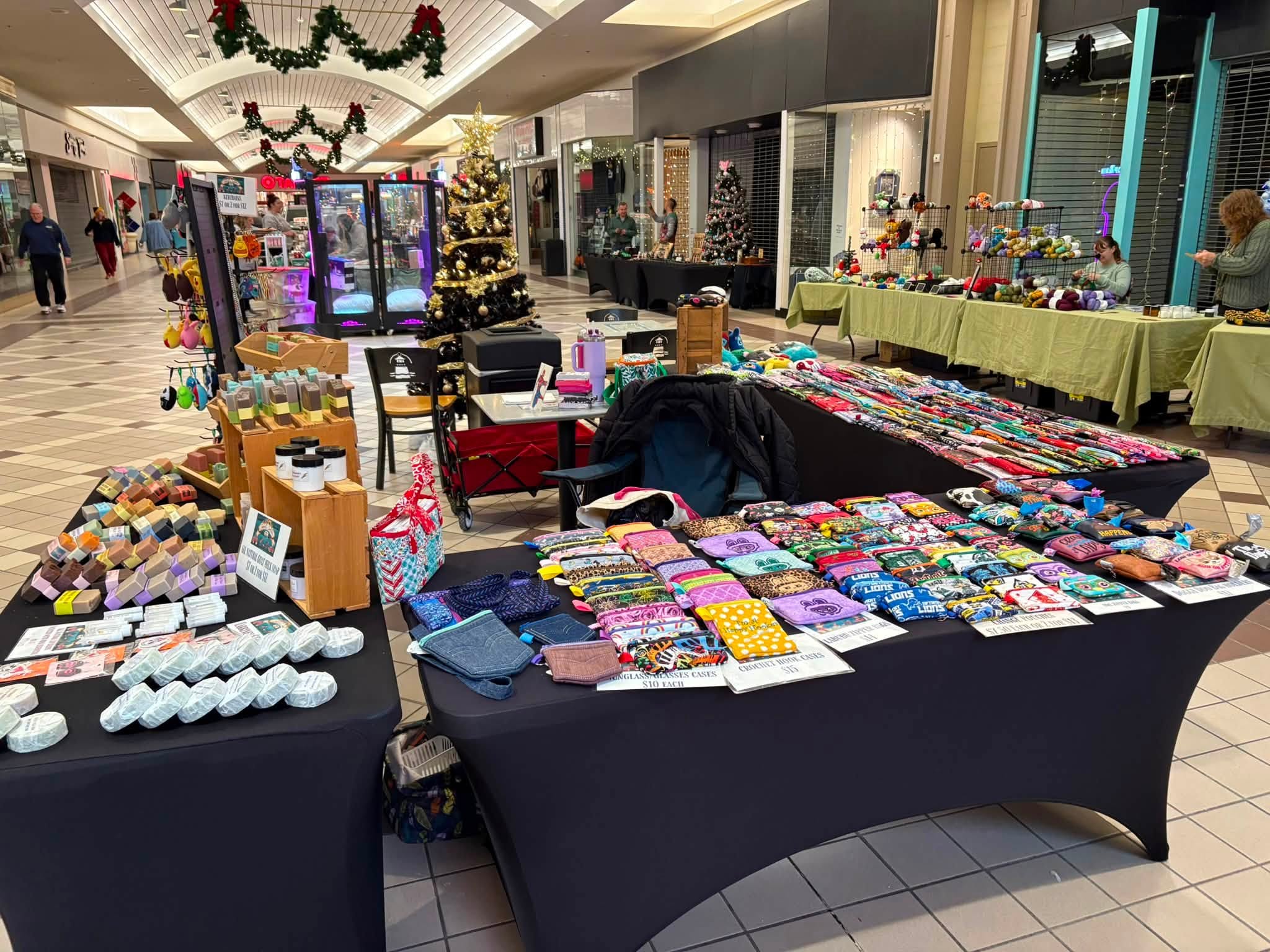 Indoor market with tables displaying colorful accessories and crafts, decorated with holiday garlands, in a shopping mall corridor.