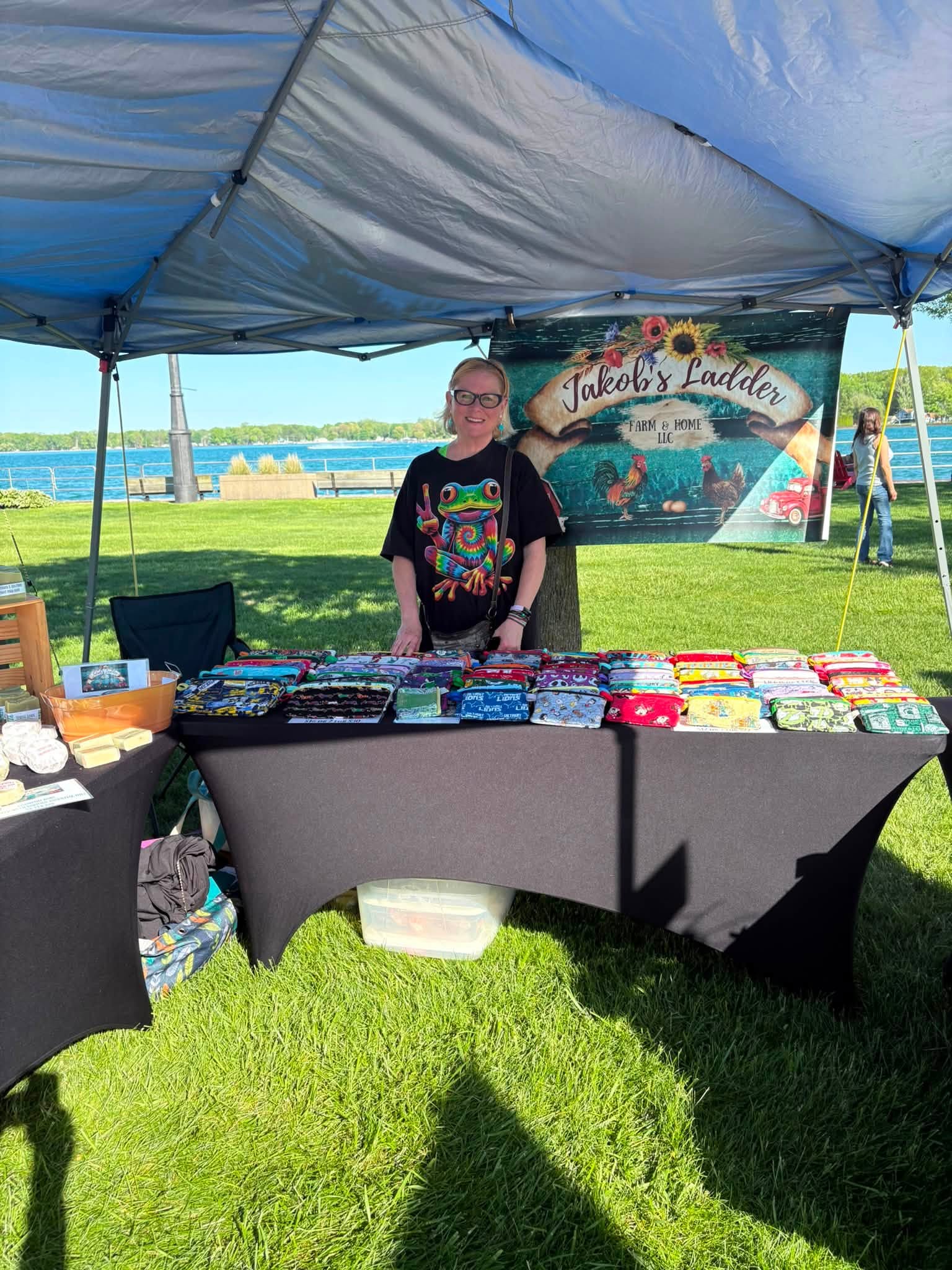 Person standing behind a table with colorful items under a tent at an outdoor event, with a banner in the background.