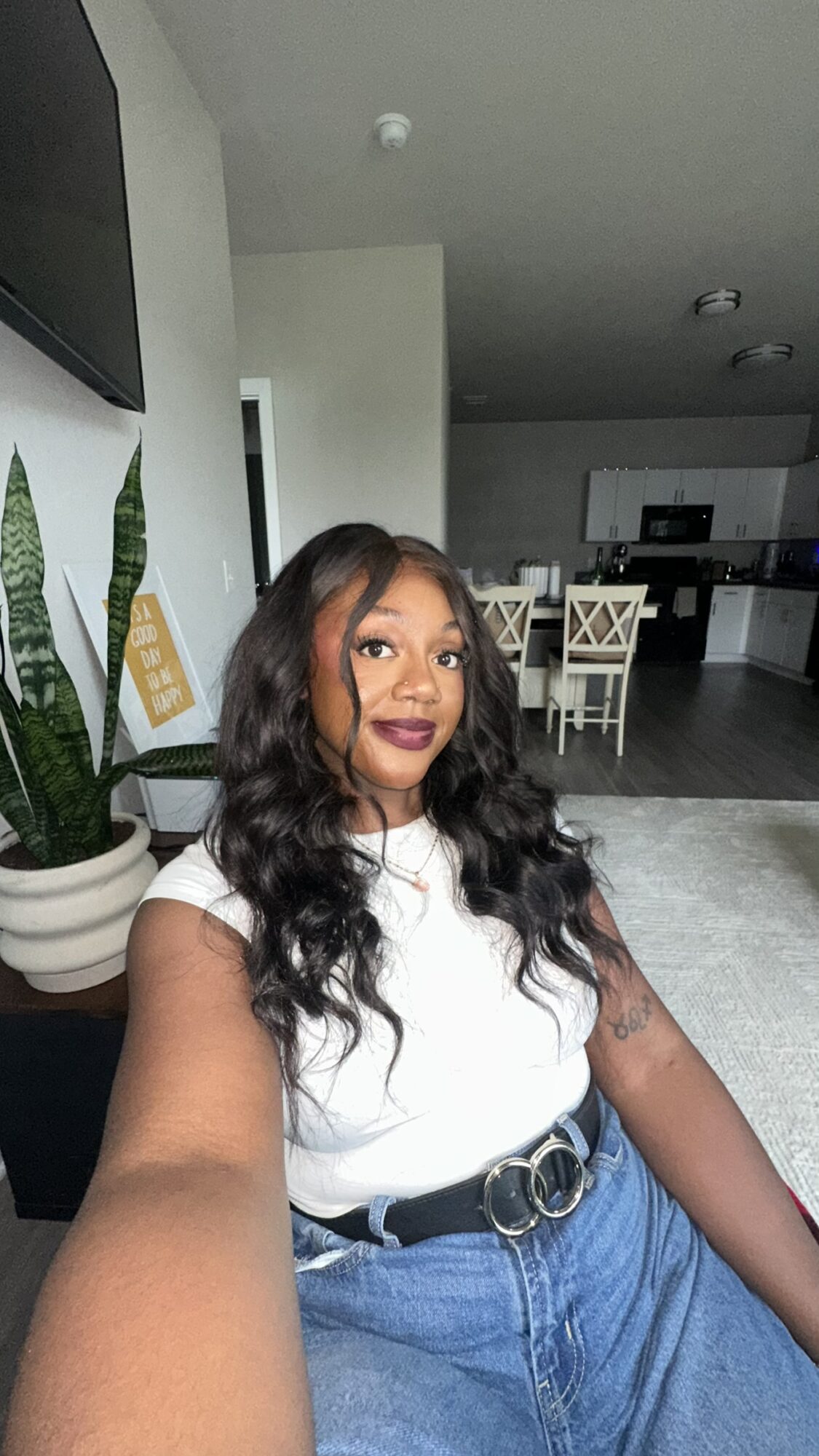 Young woman with long wavy hair taking a selfie indoors, wearing a white top and blue jeans, in a modern home.