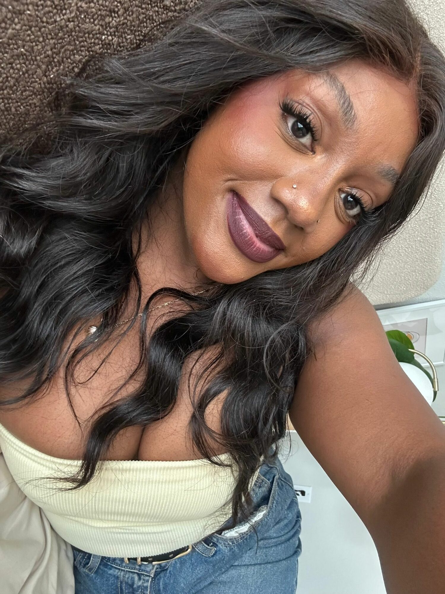 A woman with long dark hair and makeup, smiling and taking a selfie, wearing a light-colored top.