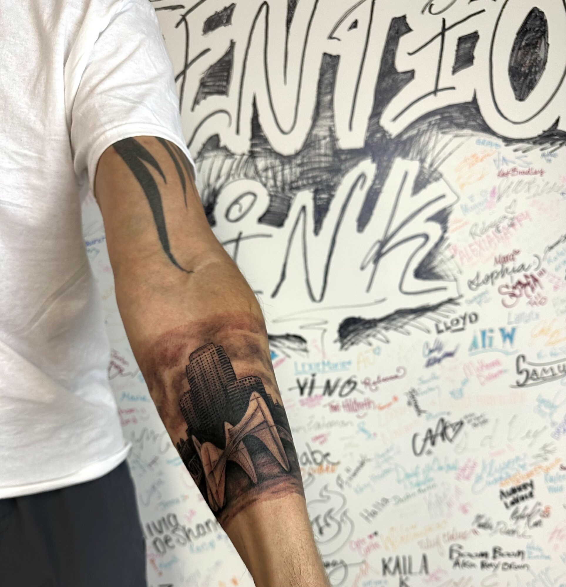 Person with a tattoo of a mountain landscape on their forearm, wearing a white shirt, standing in front of a graffiti-covered wall.