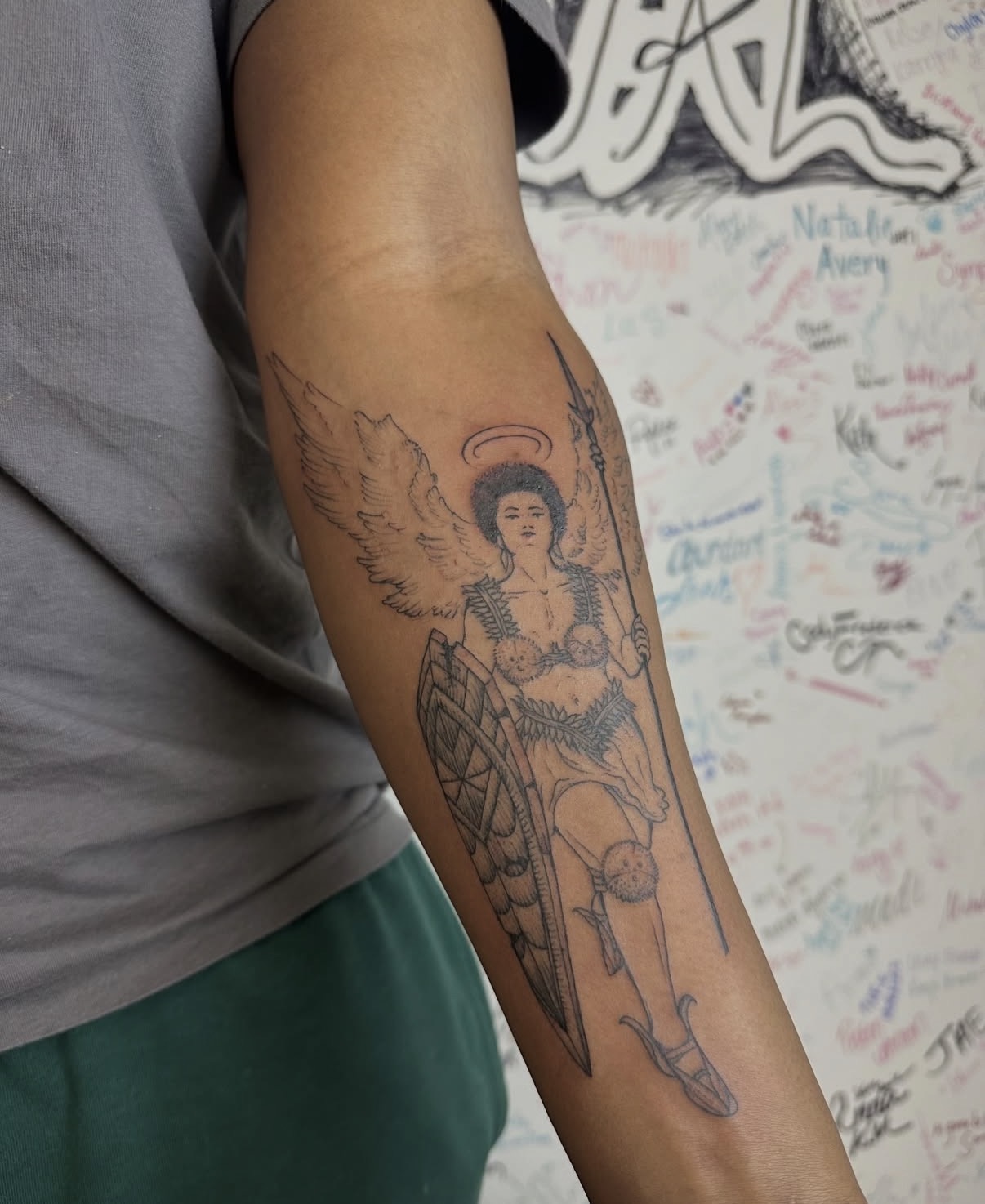 Tattoo of a seated woman with wings, holding a shield and sword, on a person's arm, with a background of signatures and writings.