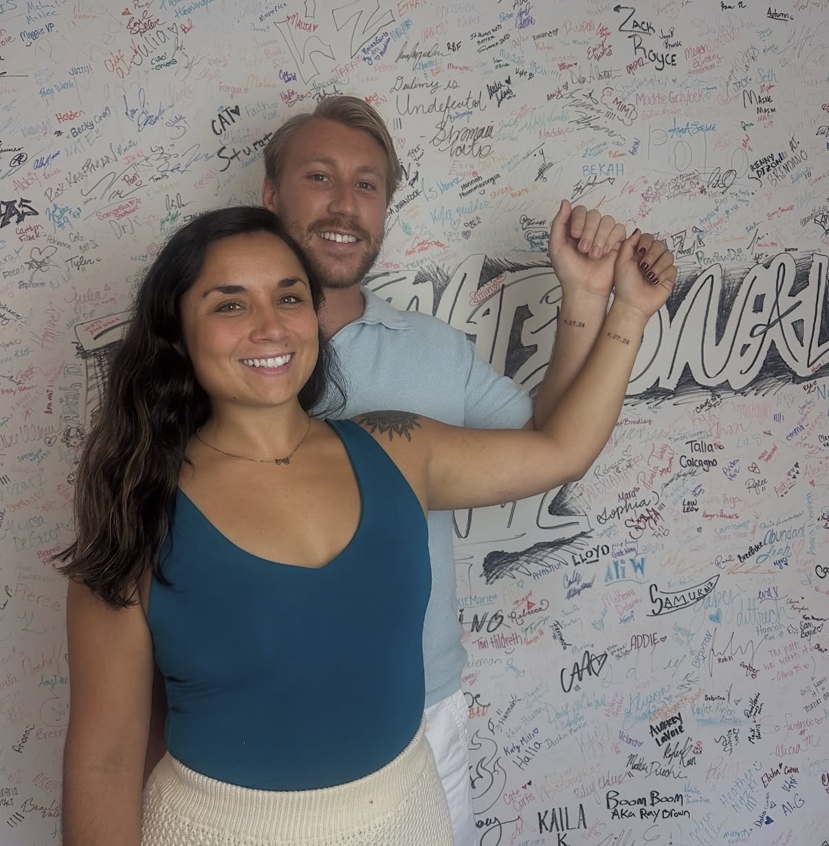 A smiling woman with long dark hair and a man with a beard, both raising their arms, standing in front of a wall with signatures.