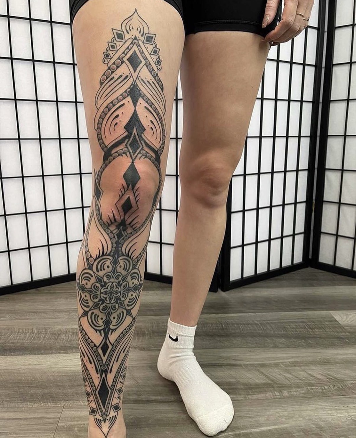 Person with a large, intricate black and gray tattoo on their leg, standing on a wooden floor with a black grid background.
