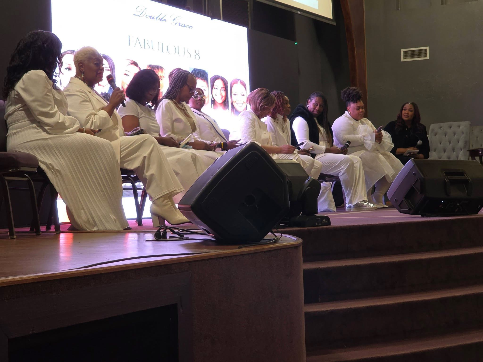 Group of people seated on stage, some holding papers, with a large screen behind them displaying text and images.