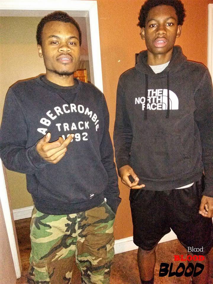 Two young men standing indoors, one making a peace sign, the other with hands in pockets, both looking at the camera.