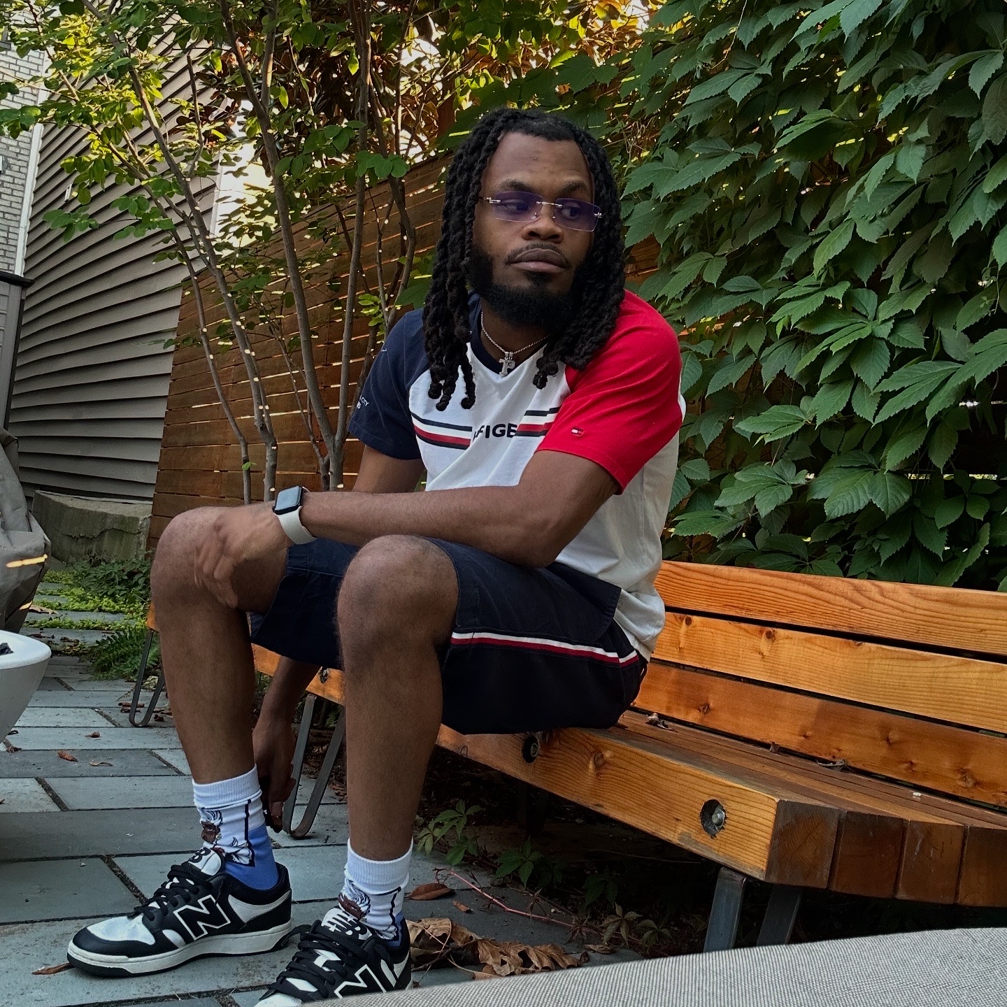 Young man with long dreadlocks, glasses, wearing a red, white, and black t-shirt, shorts, and sneakers, sitting on a wooden bench outdoors.