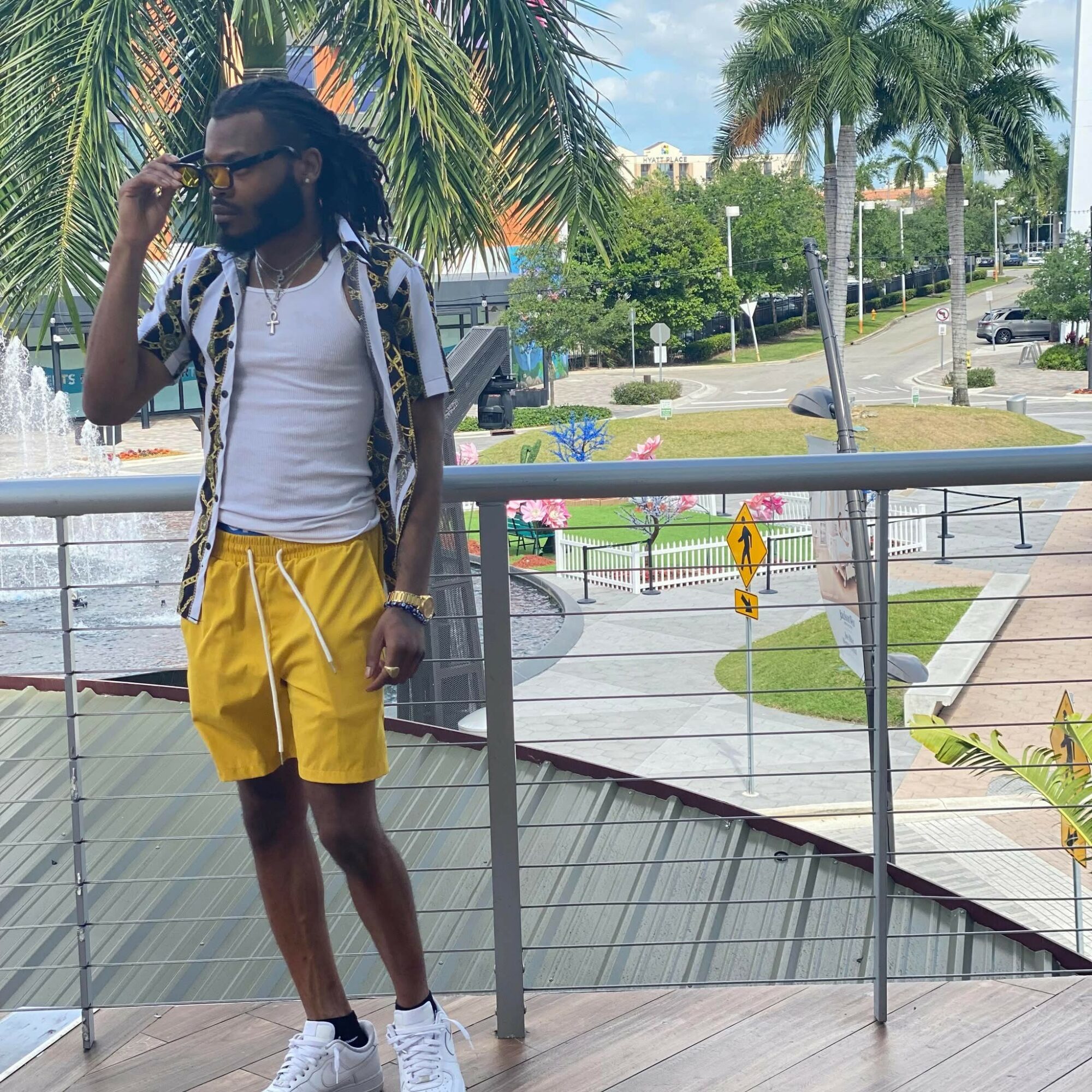 Man with sunglasses and backpack standing outdoors near palm trees and a fountain, wearing a white shirt and yellow shorts.