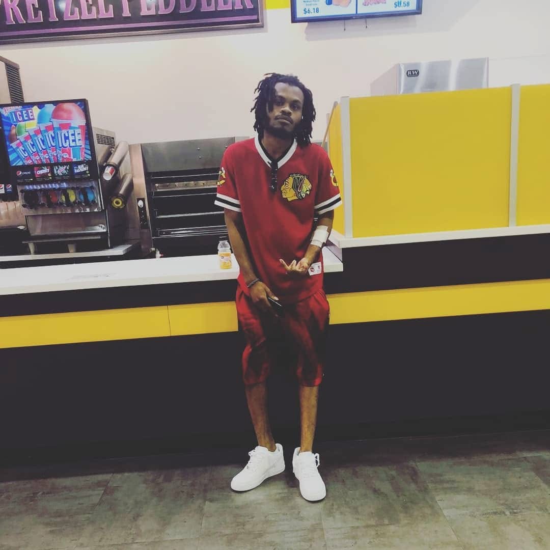 Young man with dreadlocks in a red sports jersey and shorts, standing in front of a yellow and black counter at a store.