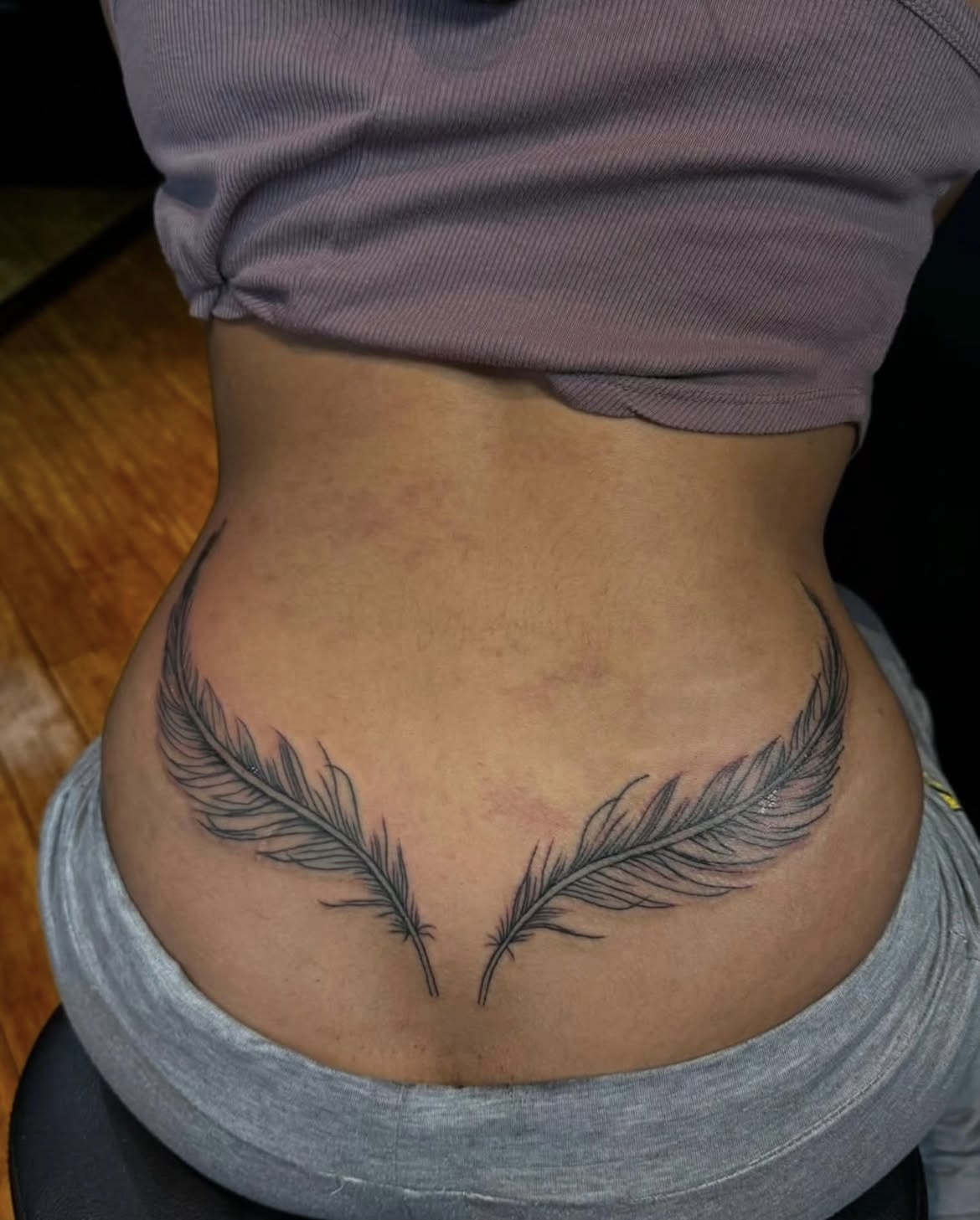Tattoo of two feathers on lower abdomen, above waistline, with a person wearing a purple top and gray pants.