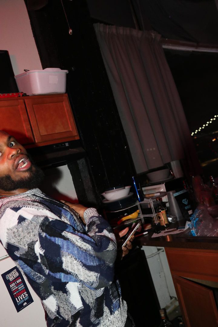 Person with a beard wearing a patterned sweater, standing in a kitchen with a window and curtains behind.