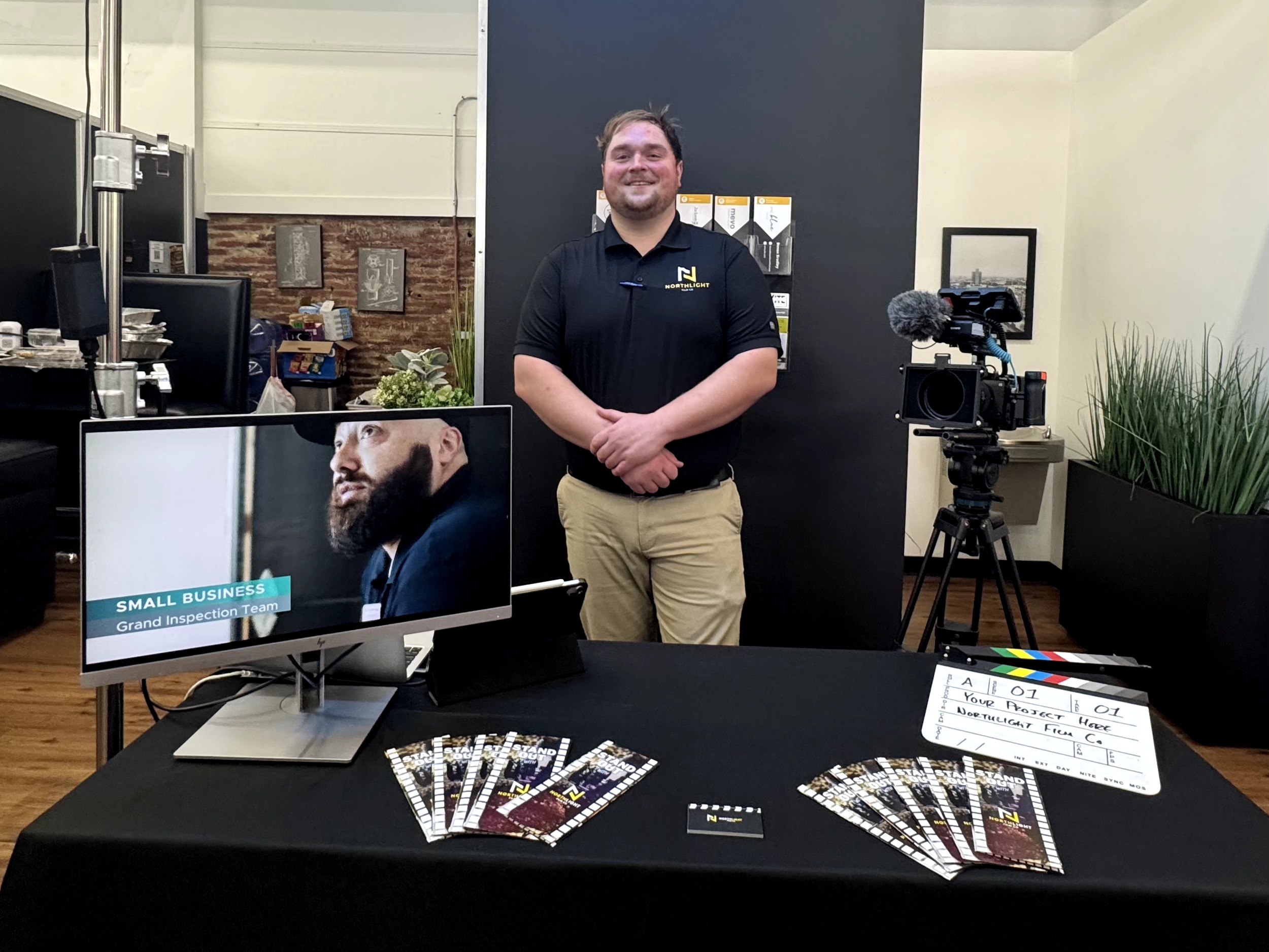 Man standing behind a table with brochures, in a room with camera equipment and a monitor, smiling at the camera.