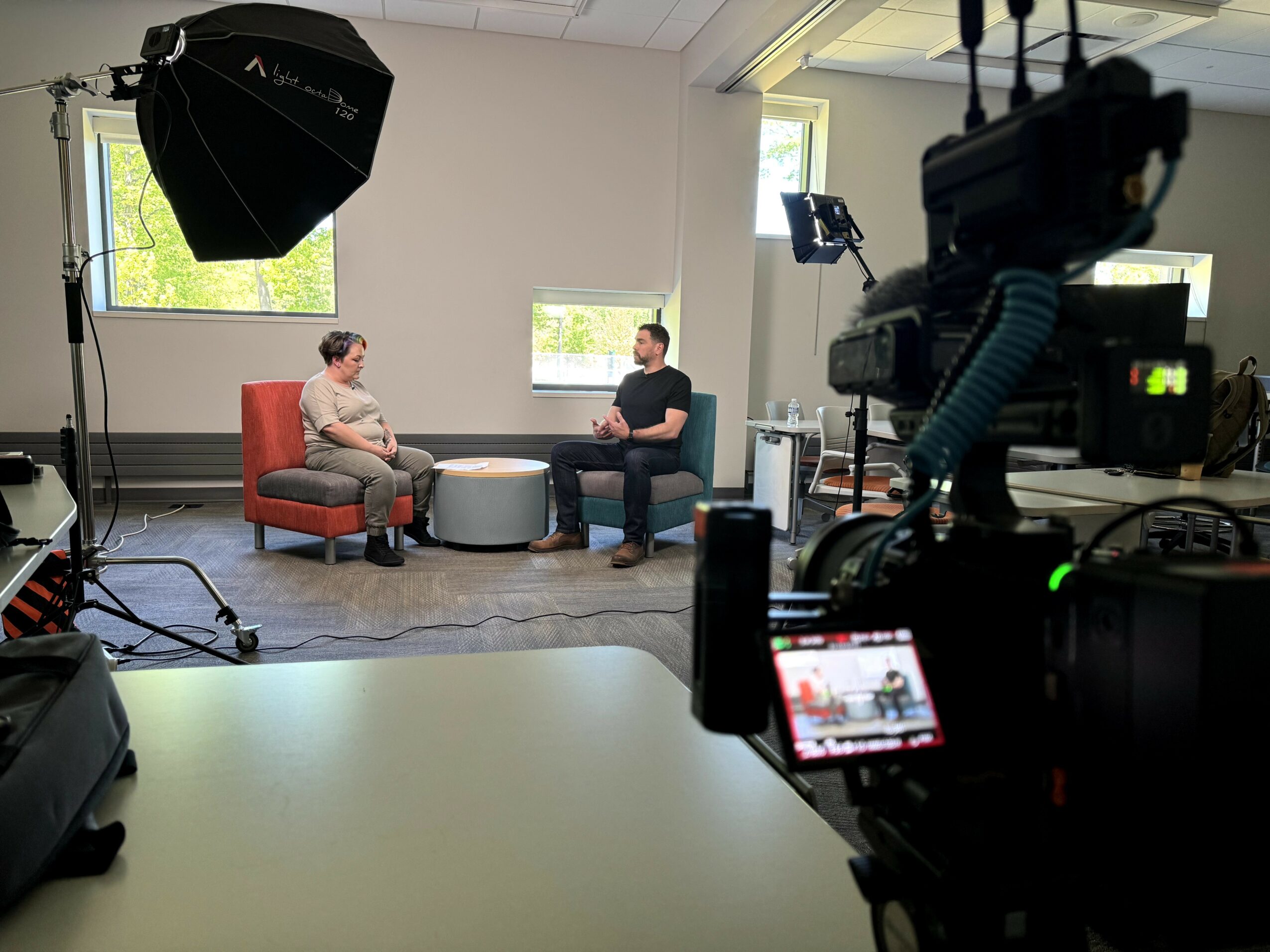 Two people sit on chairs in a studio with cameras and lighting equipment, engaged in conversation, with windows in the background.
