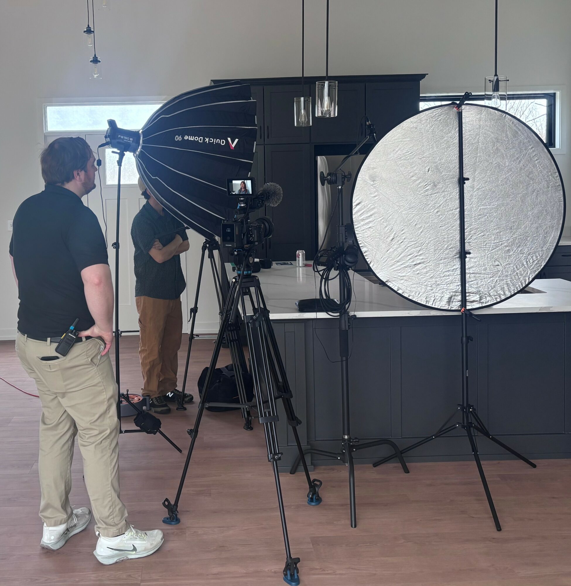 People setting up photography equipment indoors, including a large softbox and a reflector, with a window in the background.