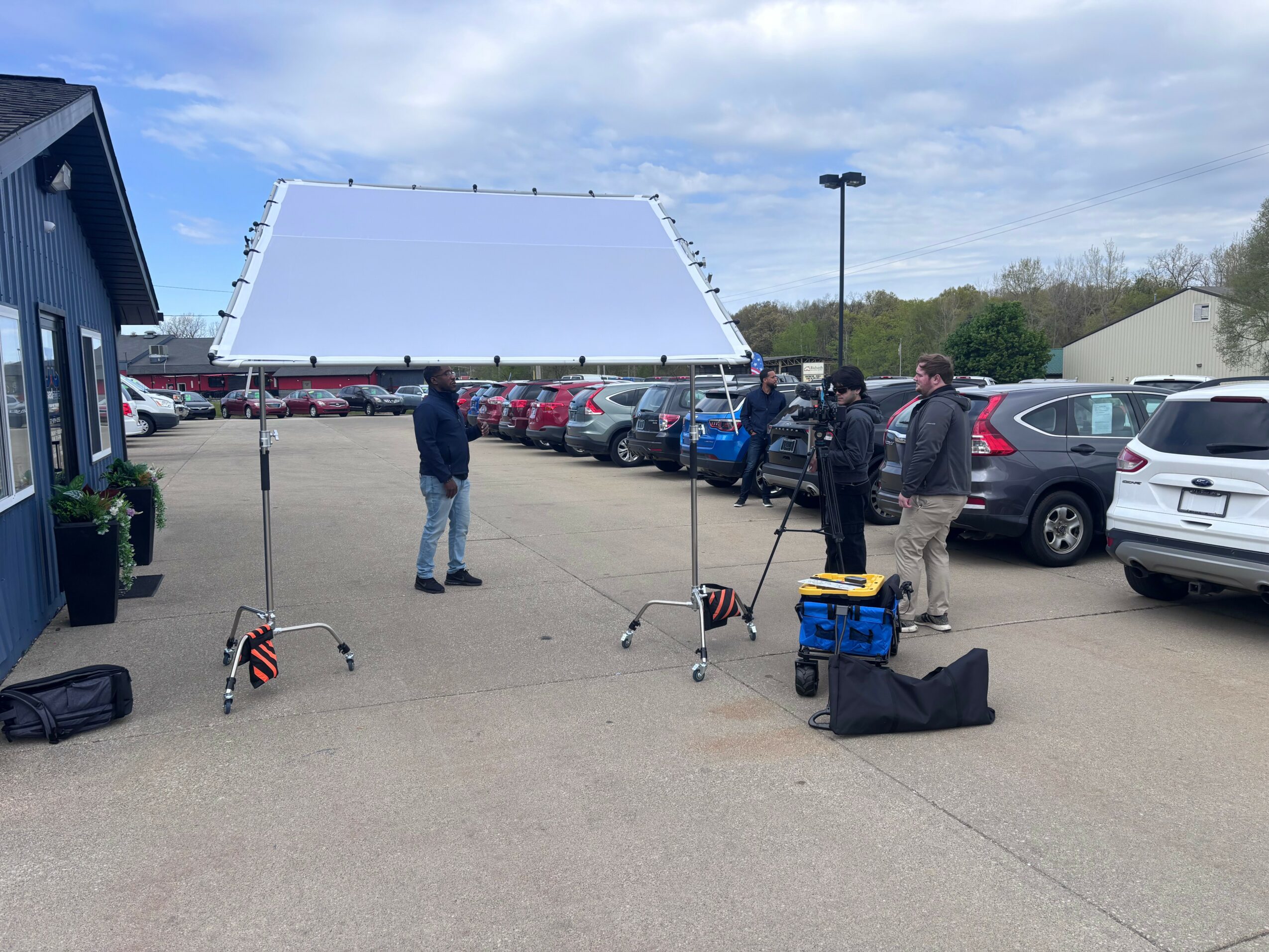 Group of people standing in a parking lot with a large white screen and equipment, cars parked nearby, blue sky with clouds.