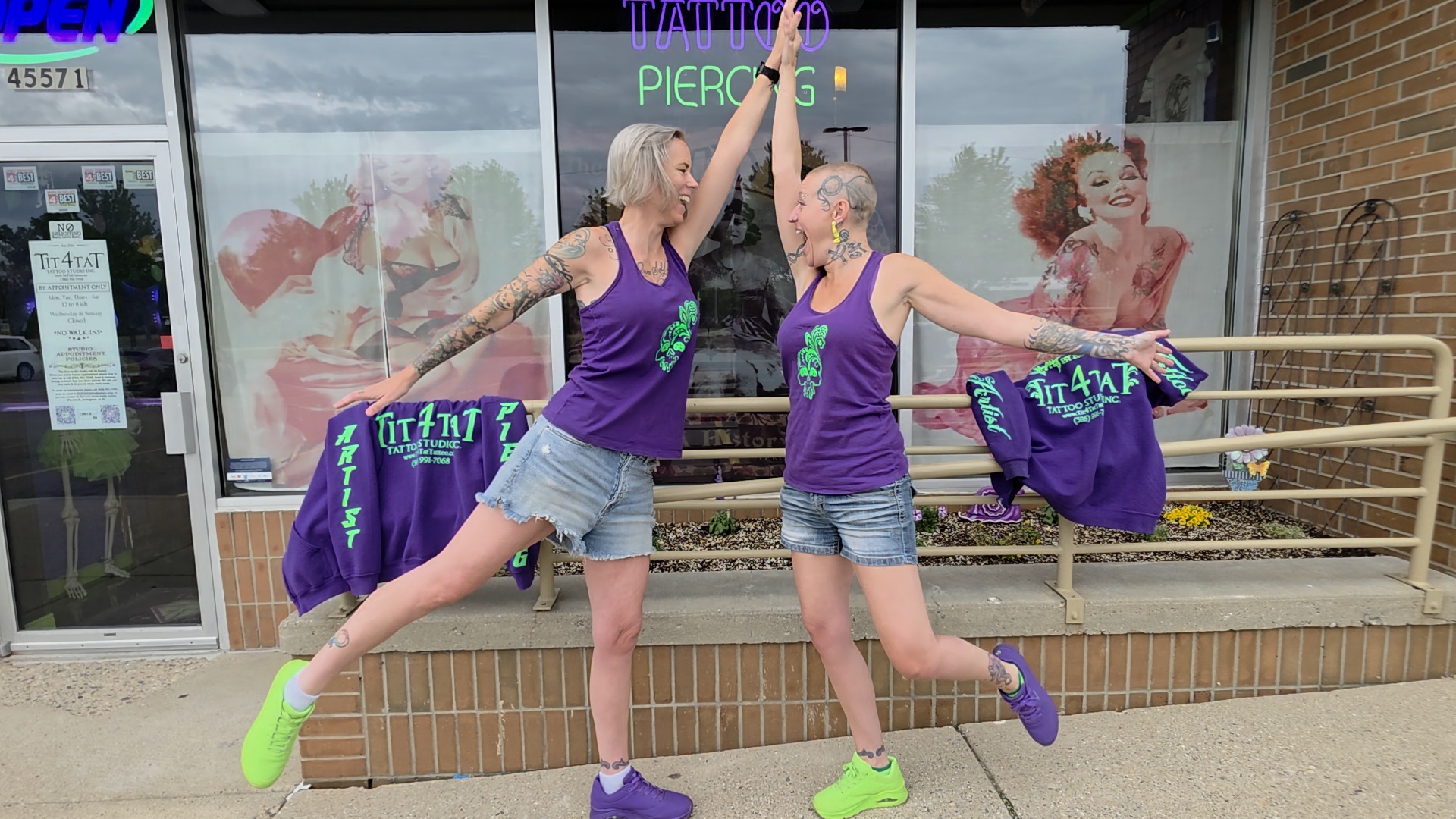 Two women with tattoos and blonde hair pose outdoors in purple tank tops and shorts, holding purple jackets, with a storefront behind them.