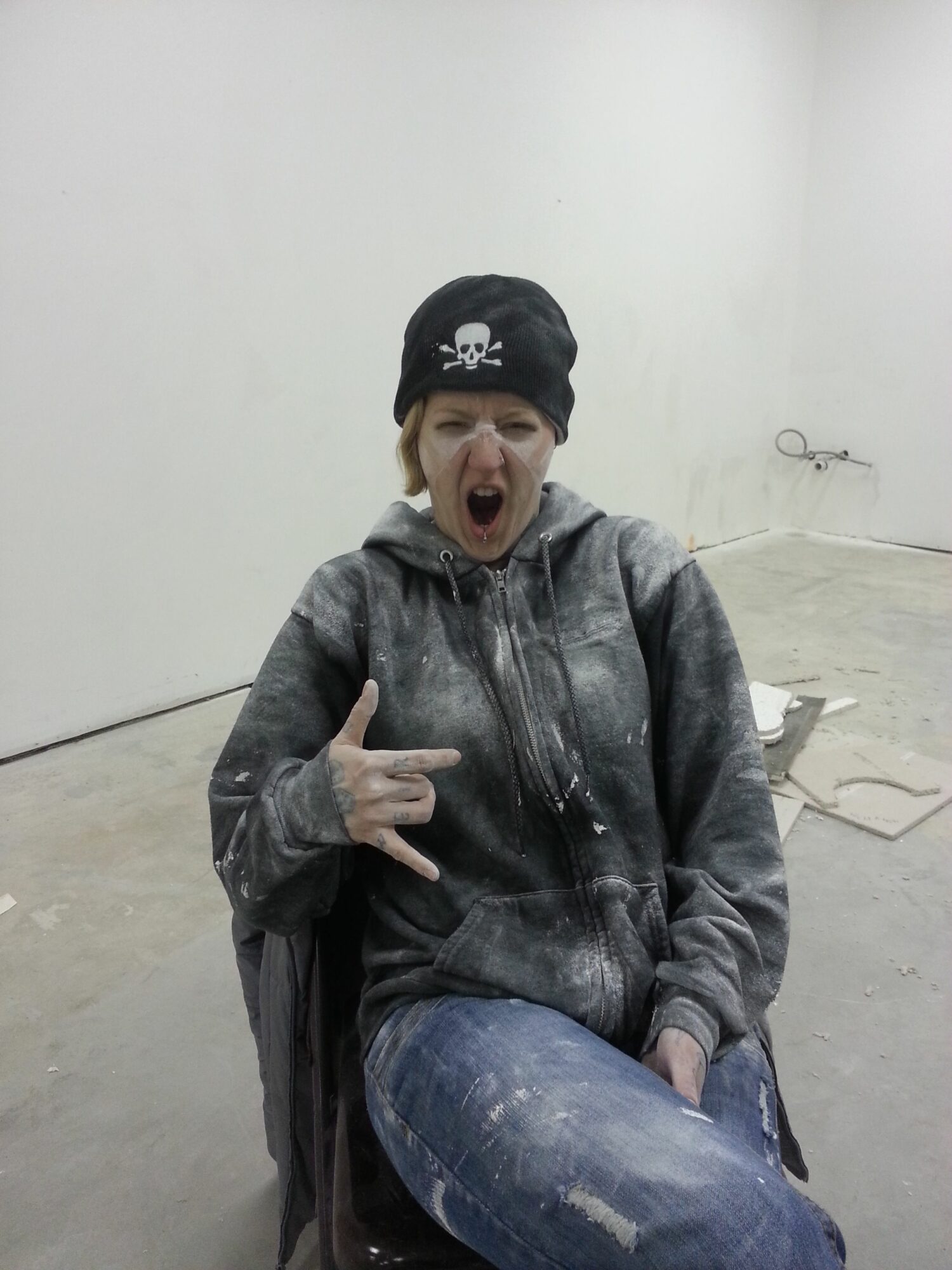 Person wearing a black beanie with a skull logo, gray jacket, and jeans, sitting on the floor with mouth open and hand gesture.