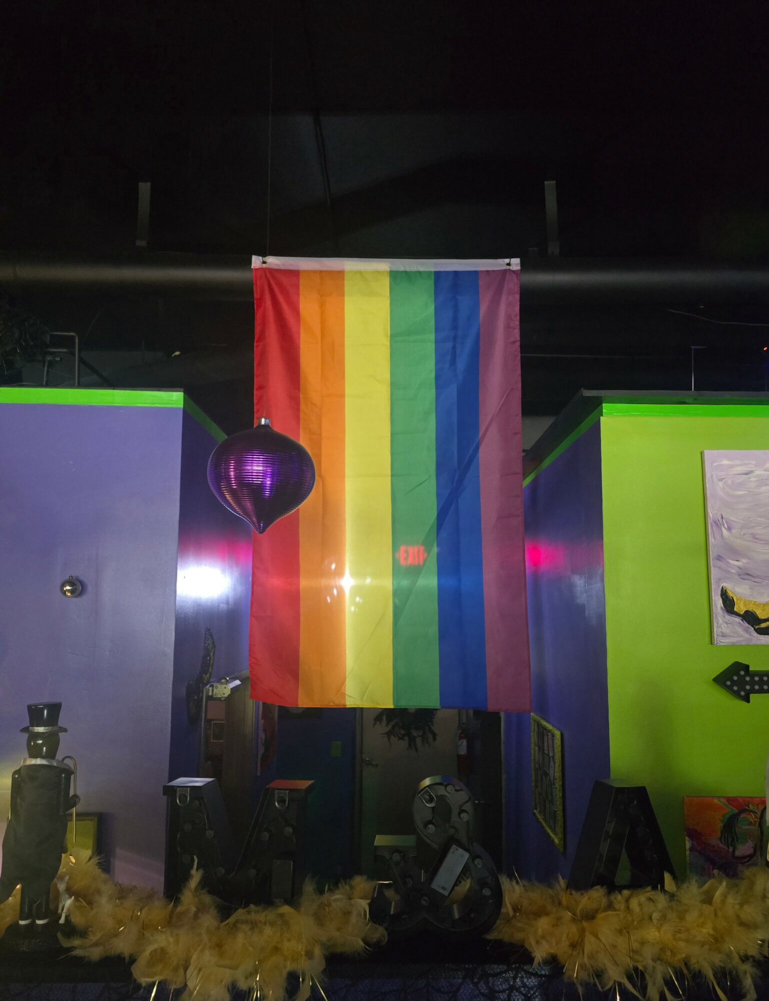 Rainbow flag hanging on a wall with purple and green walls, decorated with various objects and yellow fringe at the bottom.