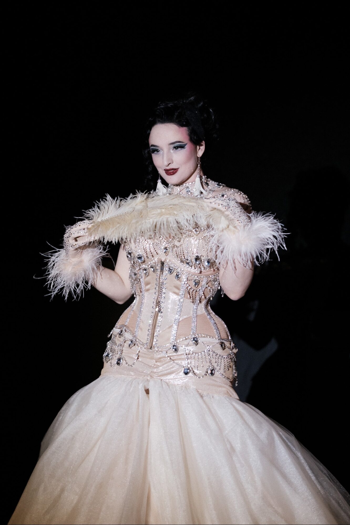 Woman in an elegant gown holding a feathered fan, standing against a dark background.