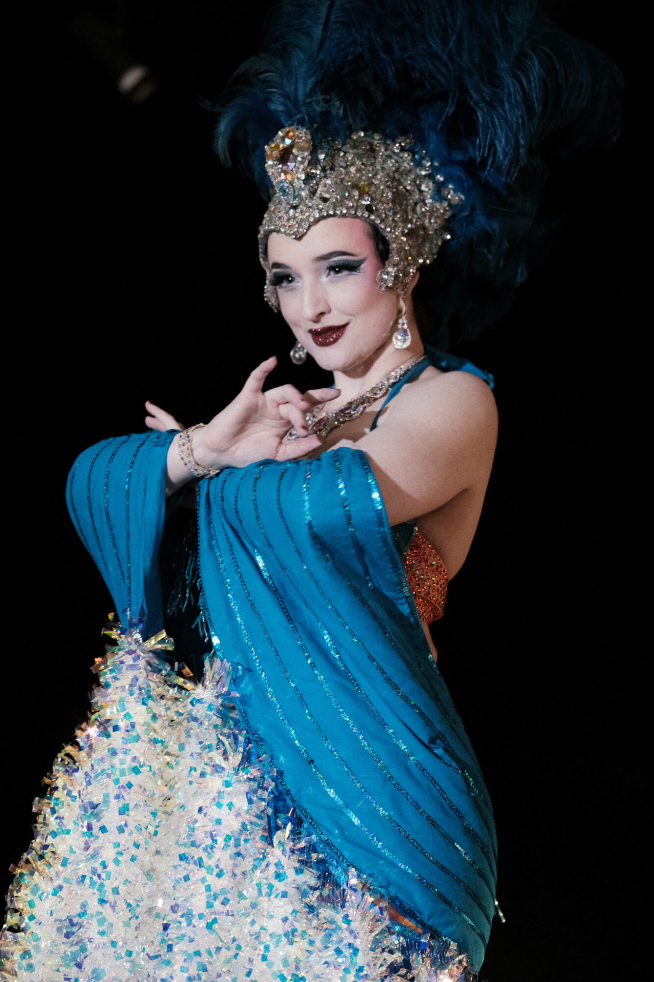 Performer in elaborate costume with blue fabric, ornate headpiece, and dramatic makeup, posing with hand near face.