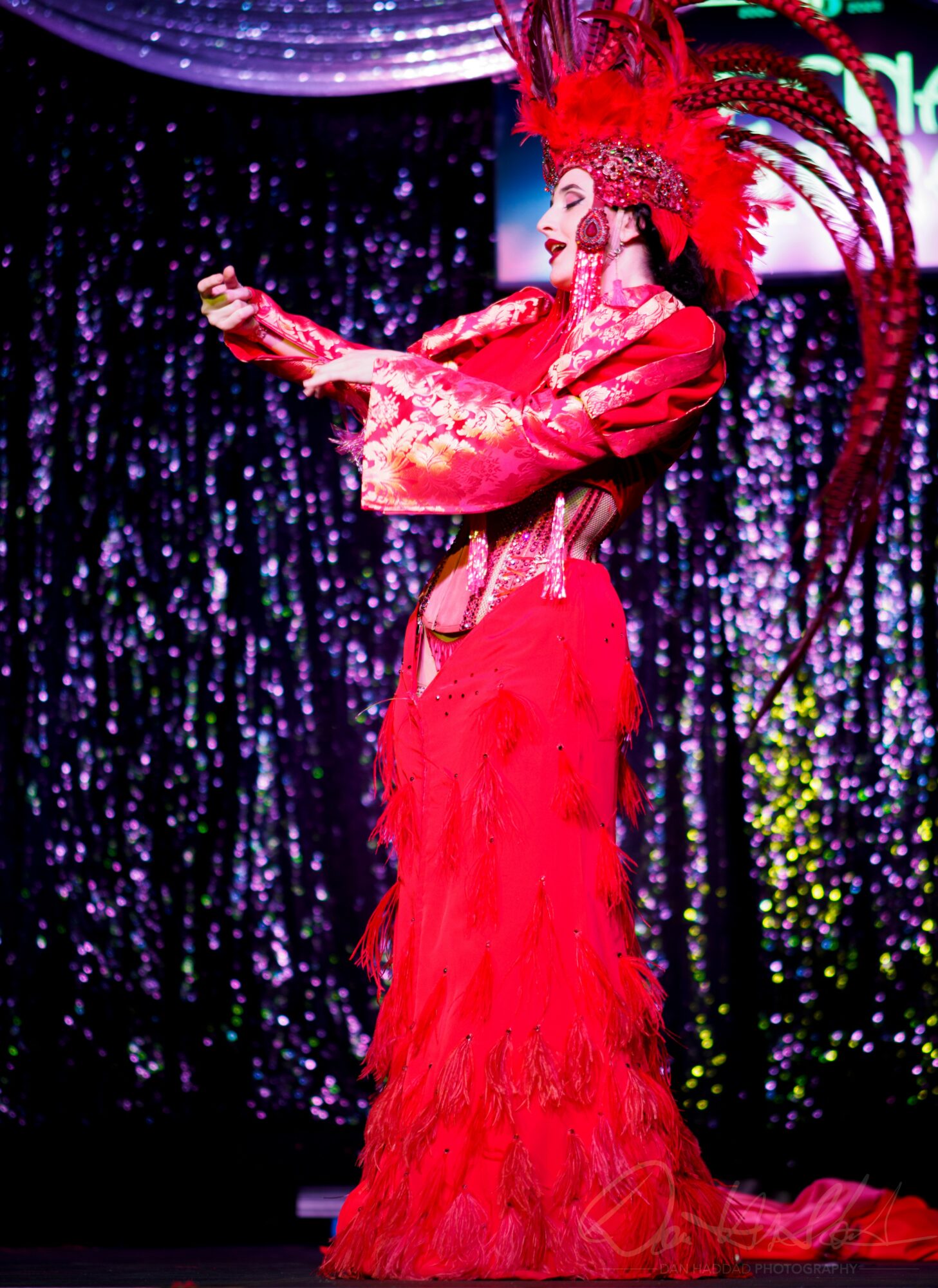 Performer in red costume with feathered headdress on stage with sparkly backdrop.