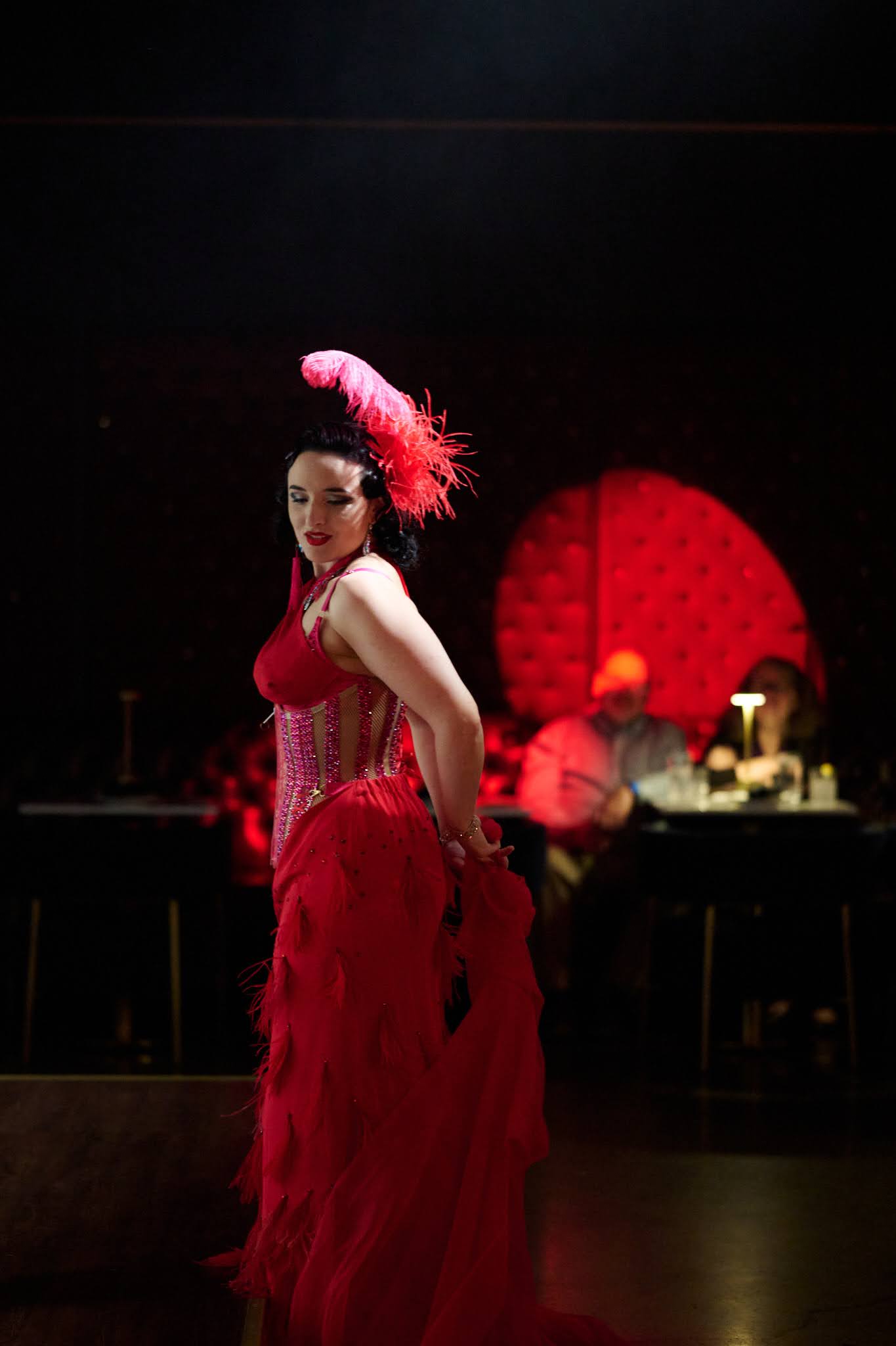 A woman in a red dress with a feathered headpiece stands on stage with a red backdrop and stage props in the background.
