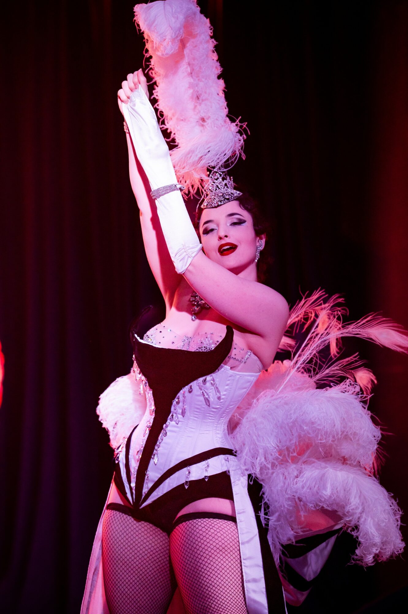 Performer in a black and white costume with feathered accessories, holding a tall feathered prop, on stage with dark background.