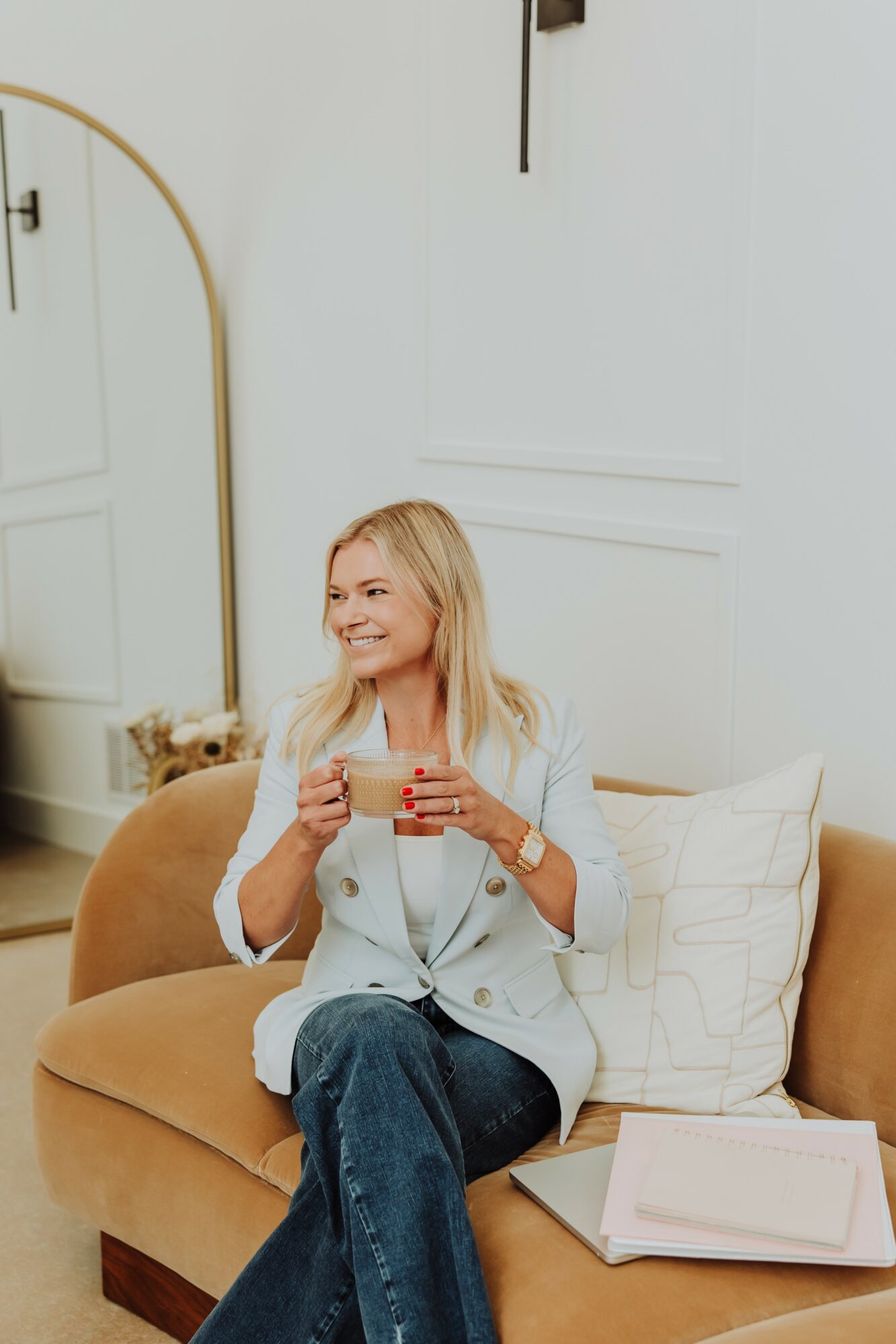 Woman with blonde hair sitting on a tan sofa, holding a red mug, smiling, in a bright room with white walls.