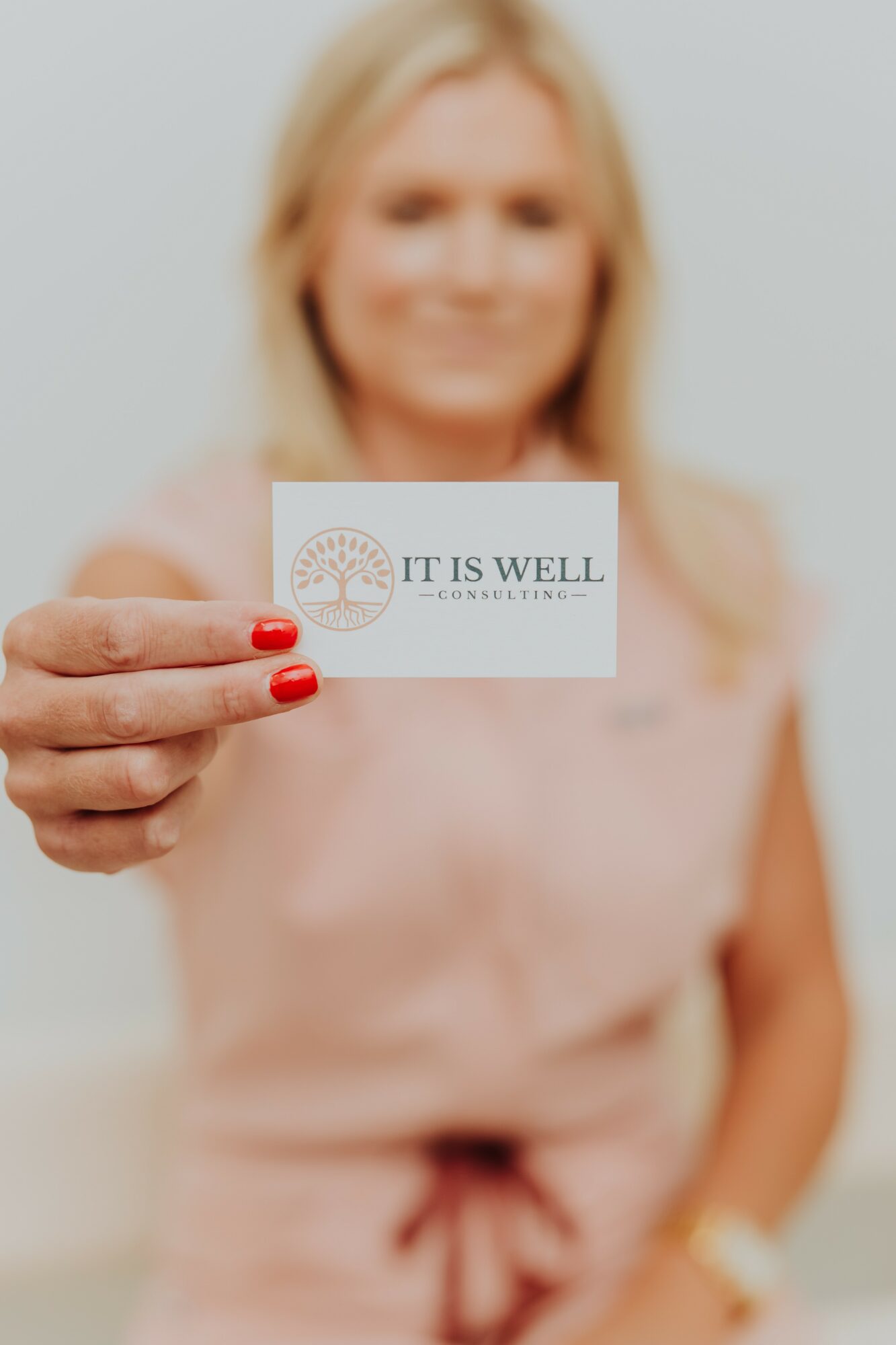 Blurry woman holding a card with text 'IT IS WELL' in front of her face, background plain light color.