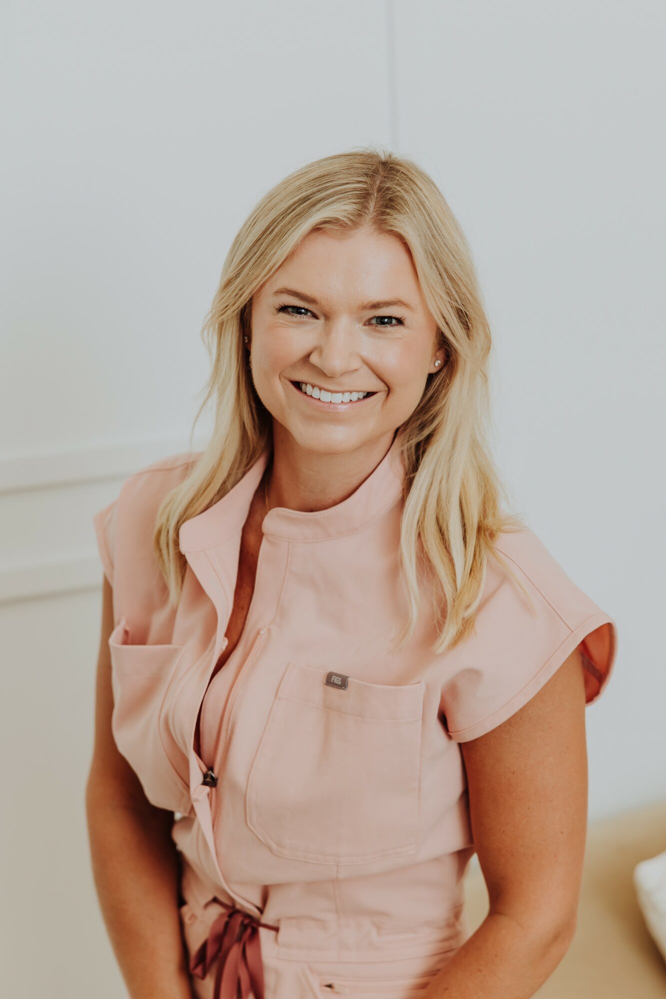 Smiling woman with blonde hair wearing a pink top, standing indoors against a light background.