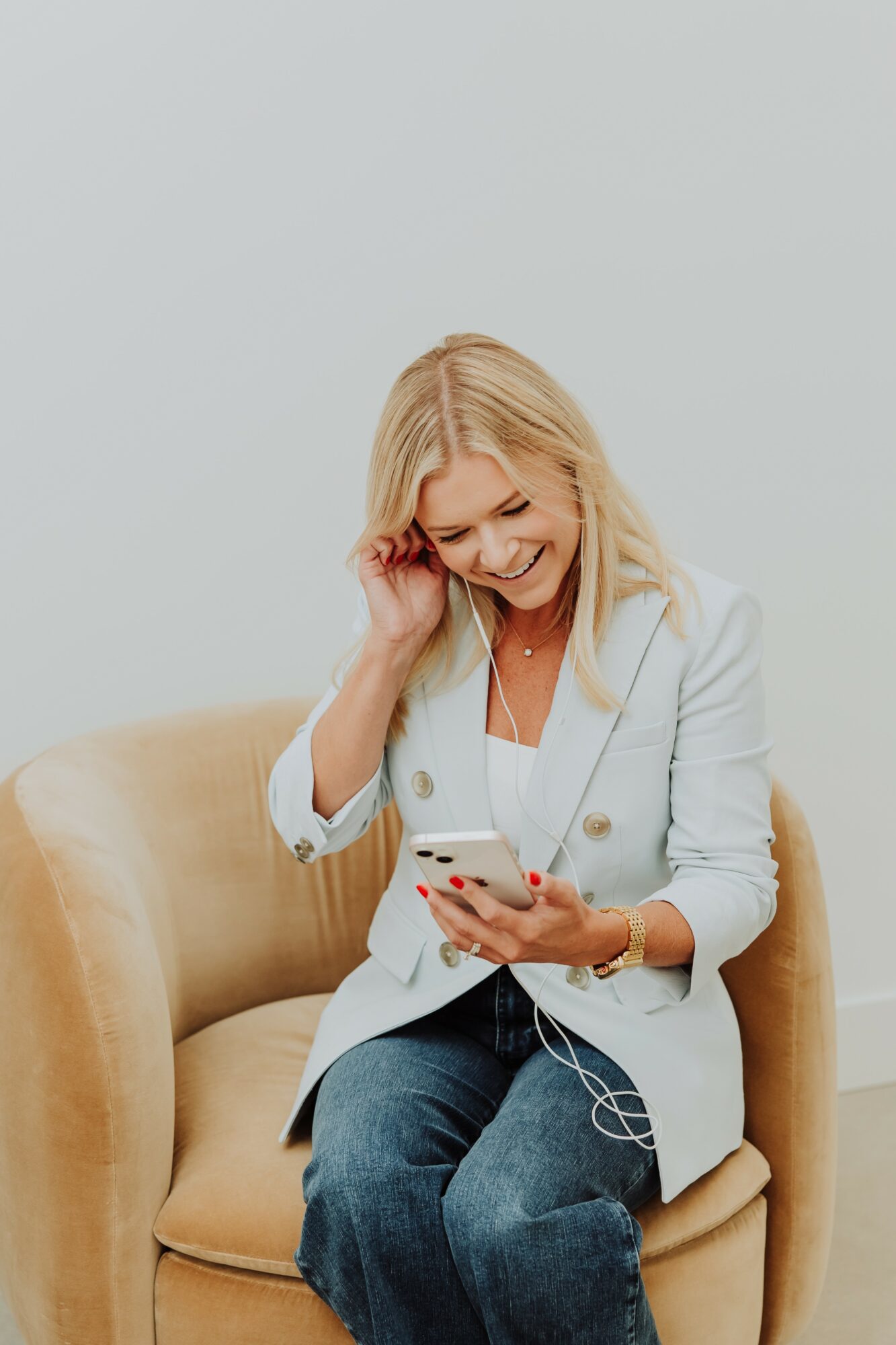 Woman with blonde hair sitting on a beige chair, smiling while looking at her phone, wearing a light blazer and jeans.